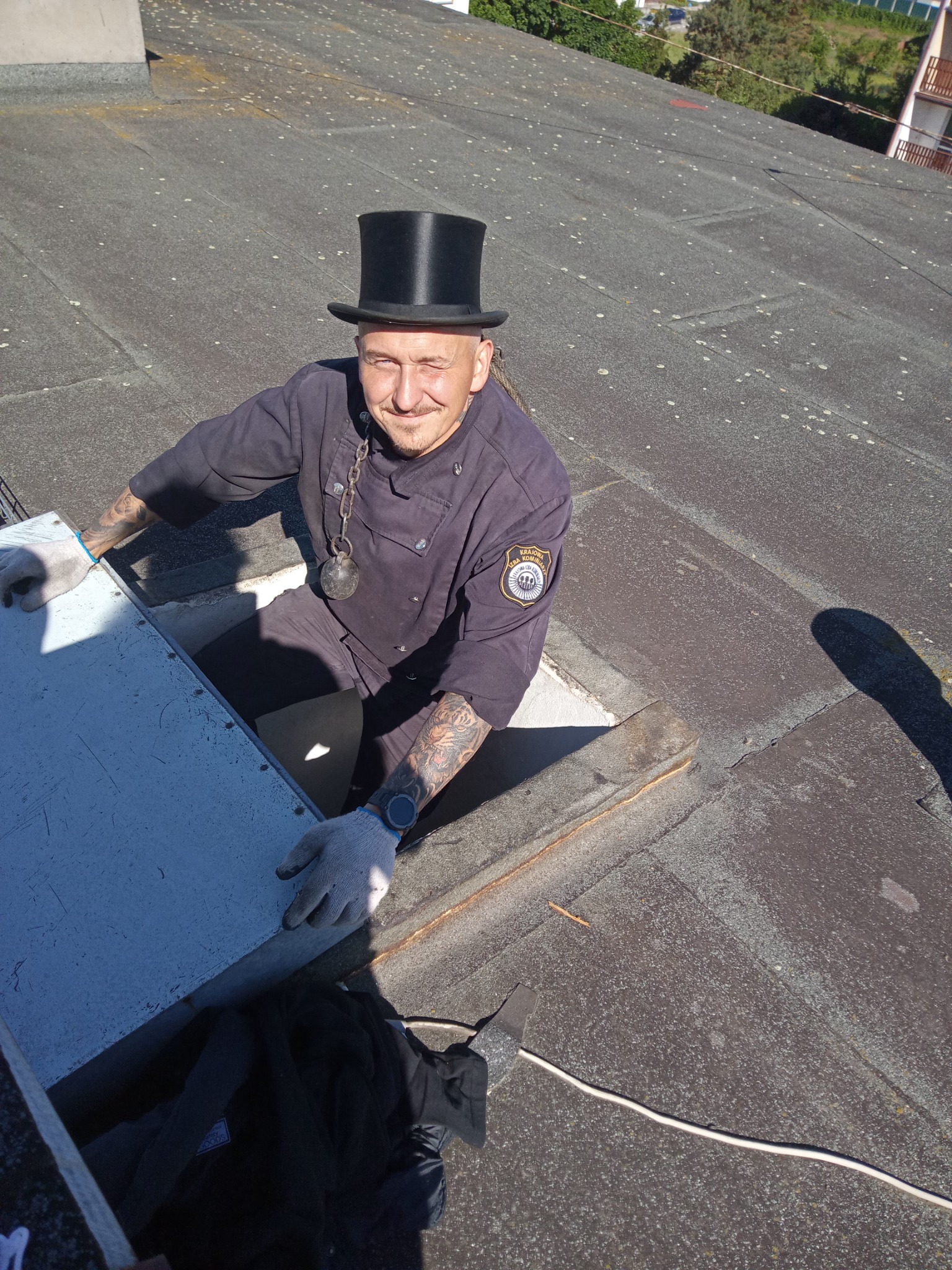 Uśmiechnięty mężczyzna w stroju roboczym i cylindrze na dachu, otwiera właz kominowy, widoczny ciężarek na łańcuchu, tatuaże na rękach i naszywka z logo firmy na ramieniu.