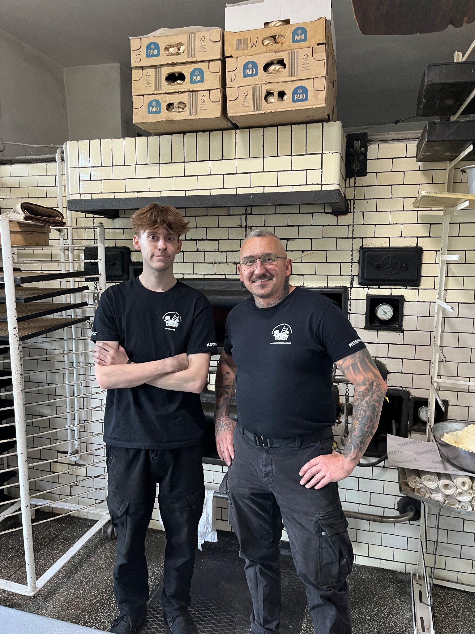 Dwóch mężczyzn w czarnych koszulkach z logo firmy kominiarskiej pozuje przed dużym, ceglanym piecem. Widoczne narzędzia i akcesoria kominiarskie.