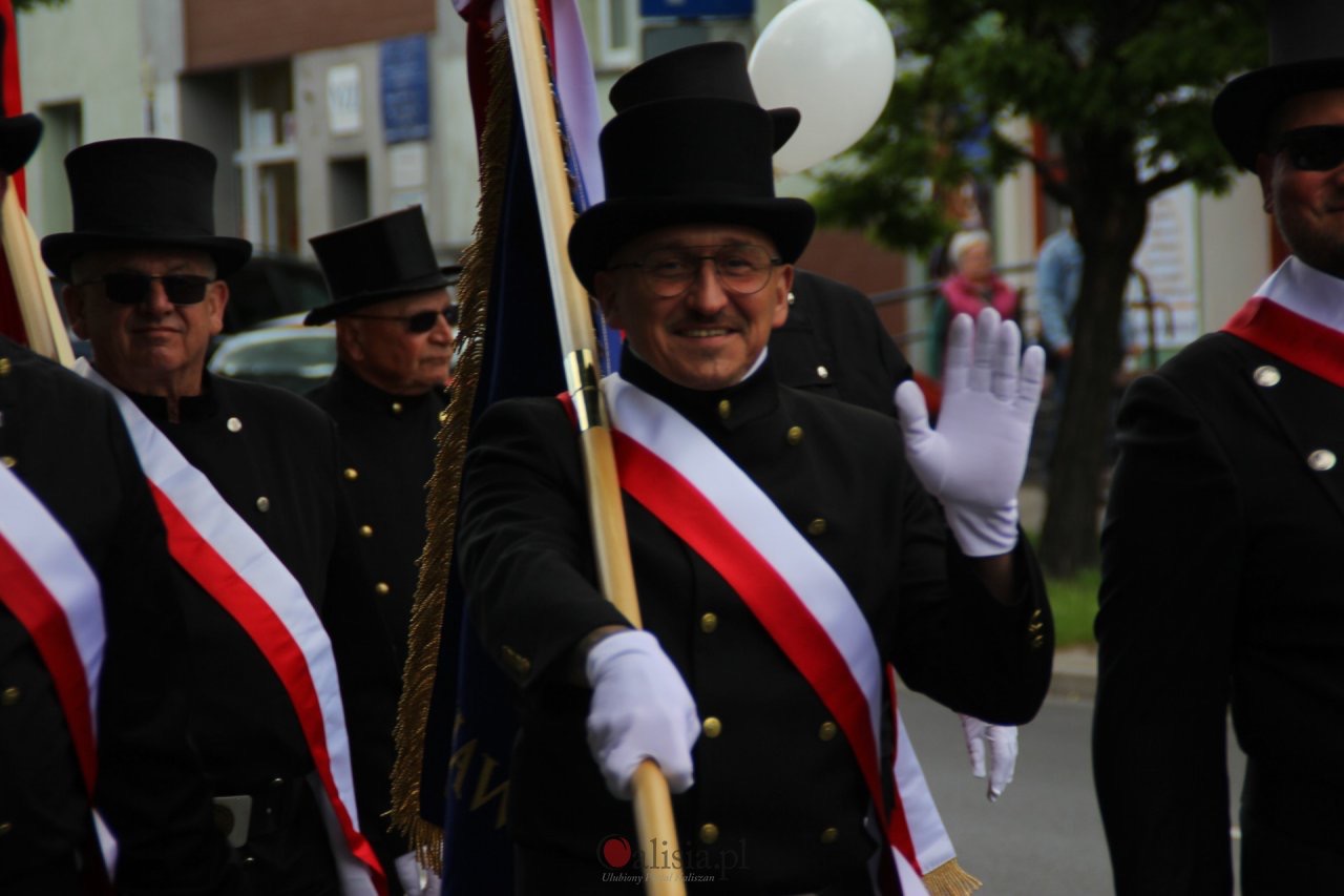 Uśmiechnięty mężczyzna w czarnym uniformie i cylindrze, z biało-czerwoną szarfą, maszeruje w pochodzie. W tle widoczni inni mężczyźni w podobnych strojach.