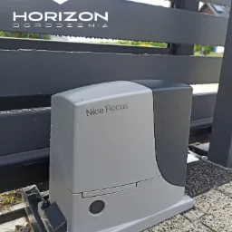 Szary automat do bram przesuwnych Nice Robus zainstalowany przy metalowym ogrodzeniu panelowym, widoczne krople deszczu.