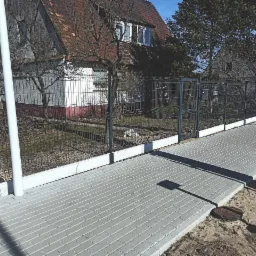 Ciemna siatka ogrodzeniowa z furtką i betonową podmurówką, widok z chodnika z szarej kostki brukowej, w tle domy jednorodzinne.