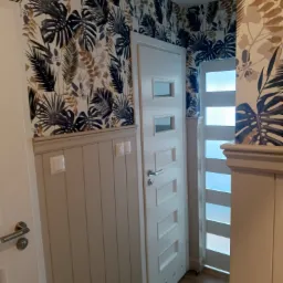 Wnętrze korytarza po remoncie: białe drzwi z przeszkleniami, lamperia w kolorze beżowym i tapeta w motywy roślinne w odcieniach brązu i granatu. Dwa reflektory na suficie.