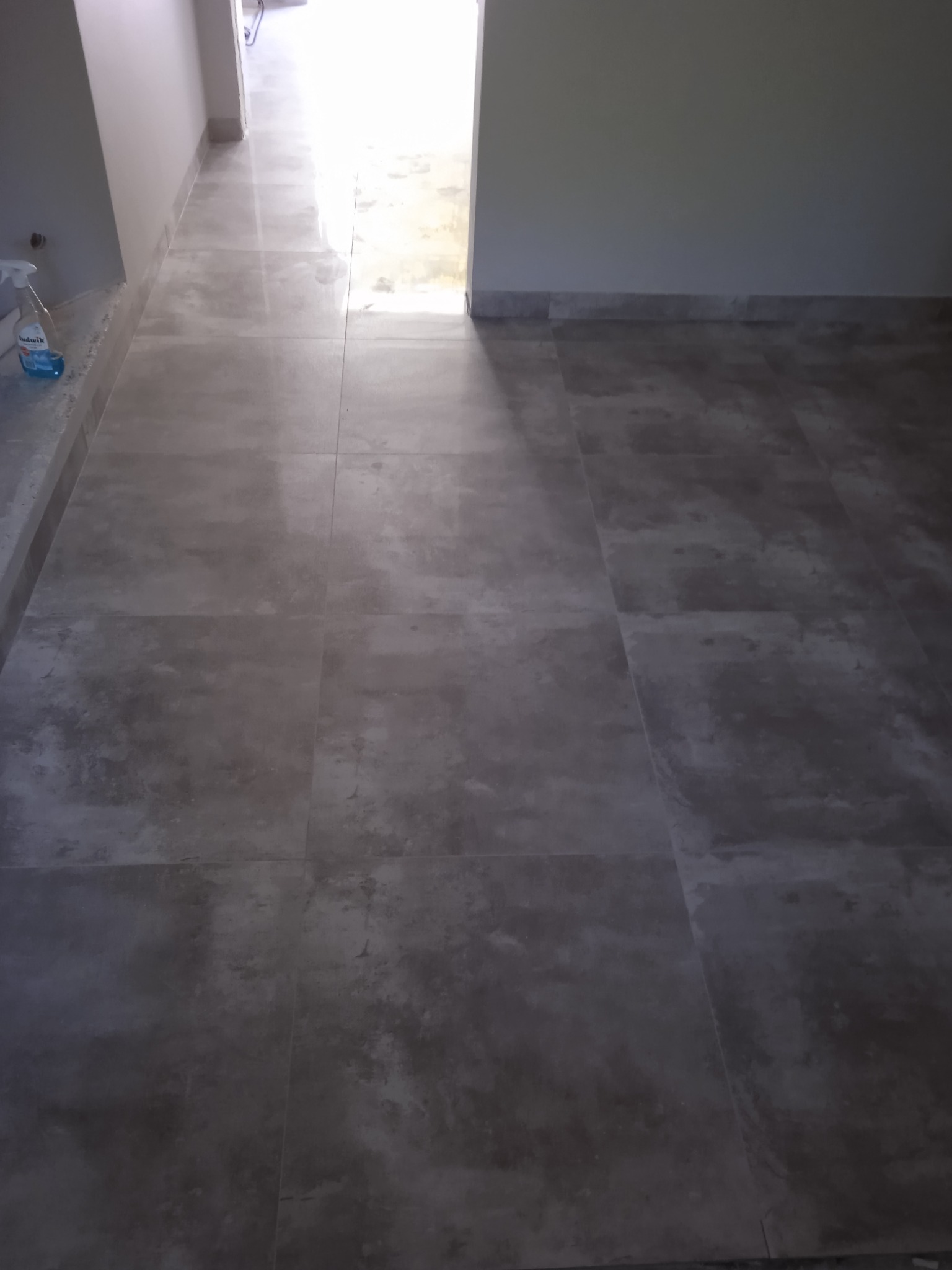 Wykończona podłoga w korytarzu wyłożona szarymi płytkami imitującymi kamień, widoczny fragment ściany z listwą przypodłogową i butelka płynu do czyszczenia w rogu.