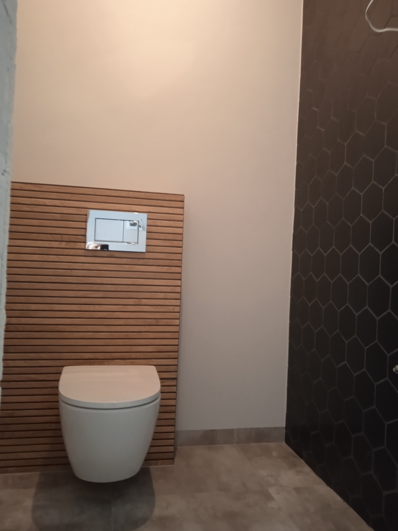 Nowoczesna toaleta podwieszana z drewnianą obudową stelaża i przyciskiem spłukującym, na ścianie ciemne heksagonalne płytki