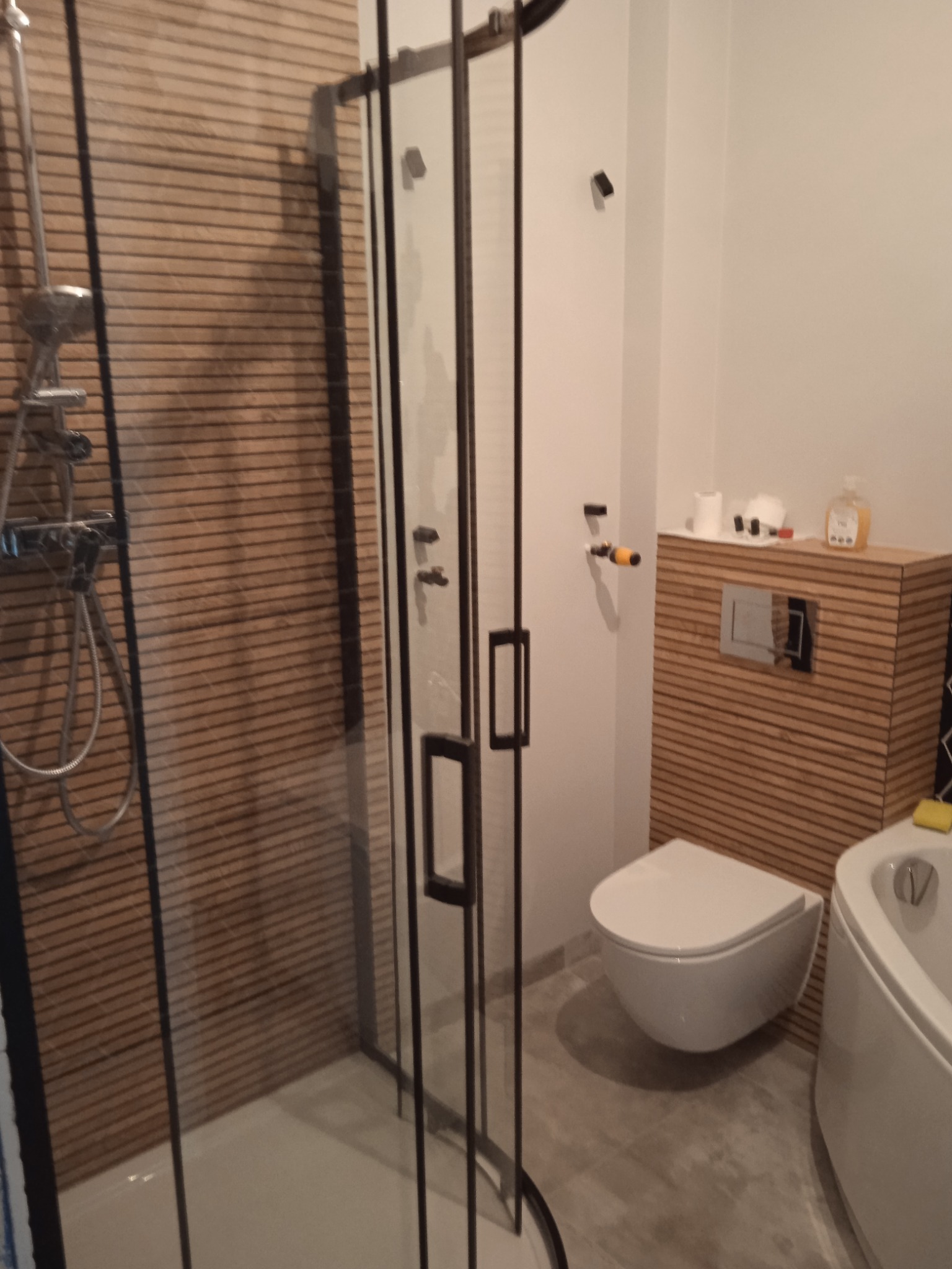 Nowoczesna łazienka z kabiną prysznicową typu walk-in z czarnymi profilami, wiszącą toaletą z drewnianą obudową i baterią podtynkową oraz wanną z hydromasażem.