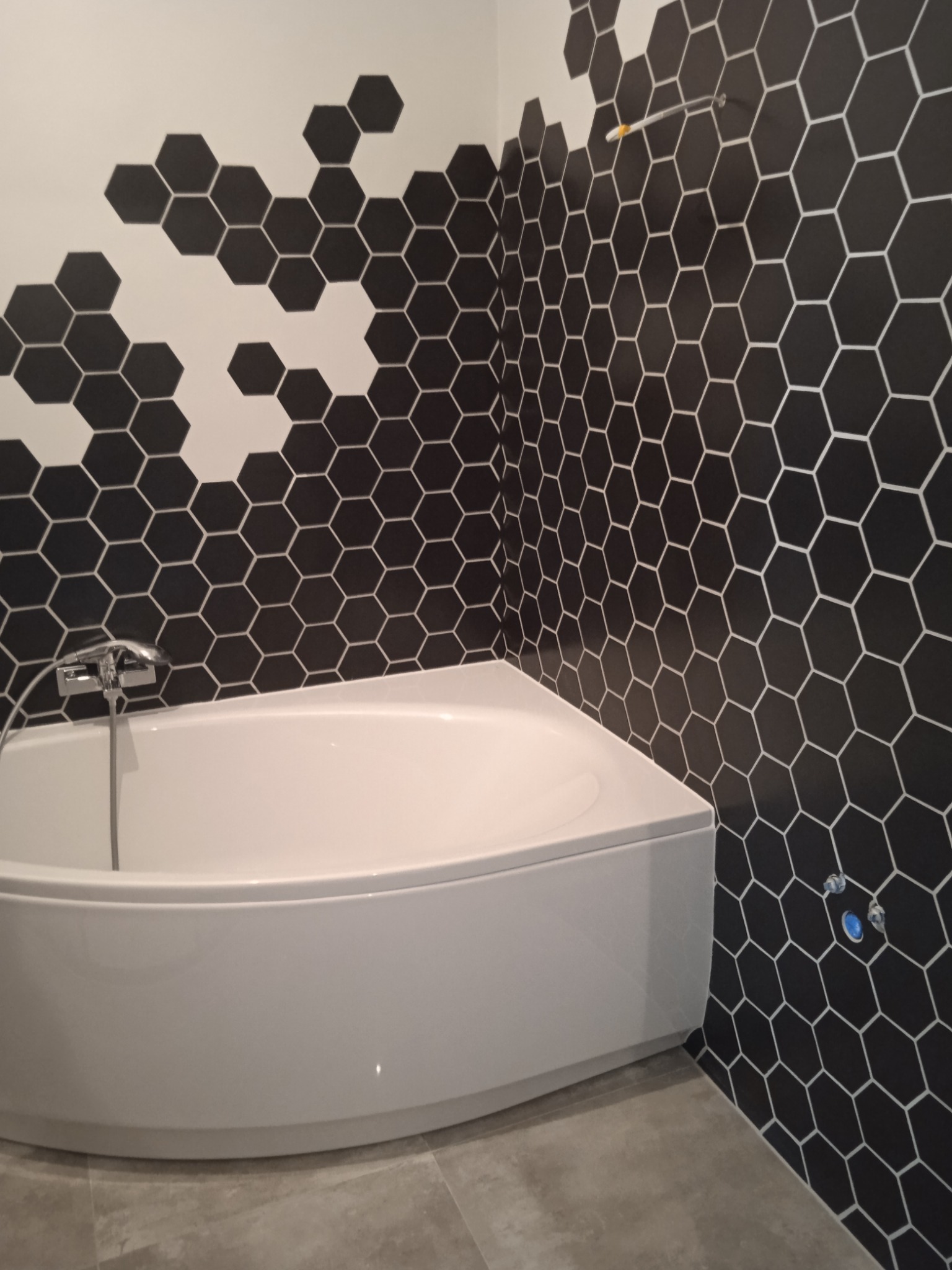 Nowoczesna łazienka z białą, narożną wanną i ścianą wyłożoną czarnymi, heksagonalnymi płytkami z białymi fugami, tworzącymi efektowny wzór z przejściem do białych płytek w górnej części ściany.