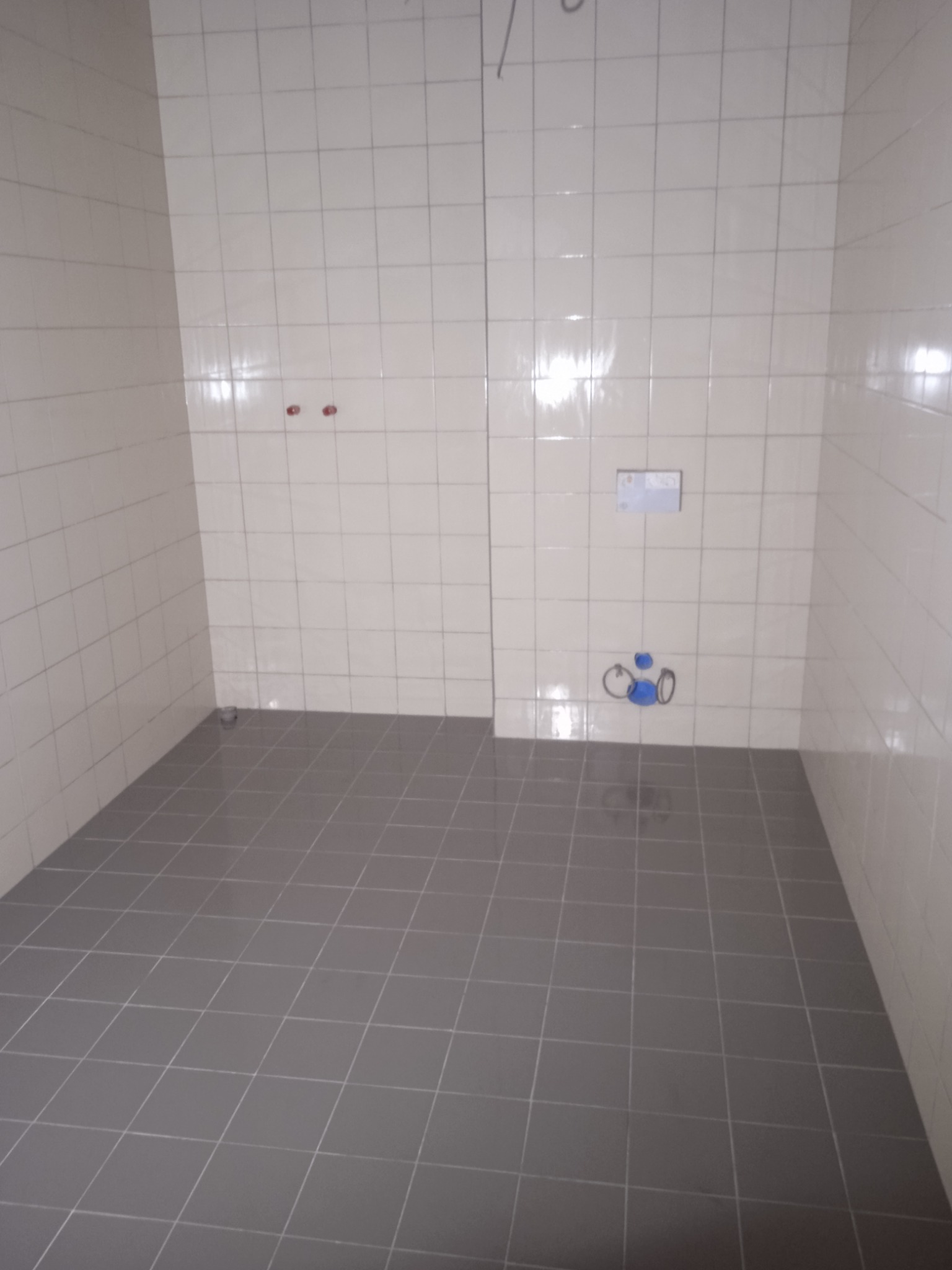 Pusta łazienka w trakcie wykańczania, z szarymi płytkami podłogowymi i białymi płytkami ściennymi, widoczne przyłącza wodne i kanalizacyjne.