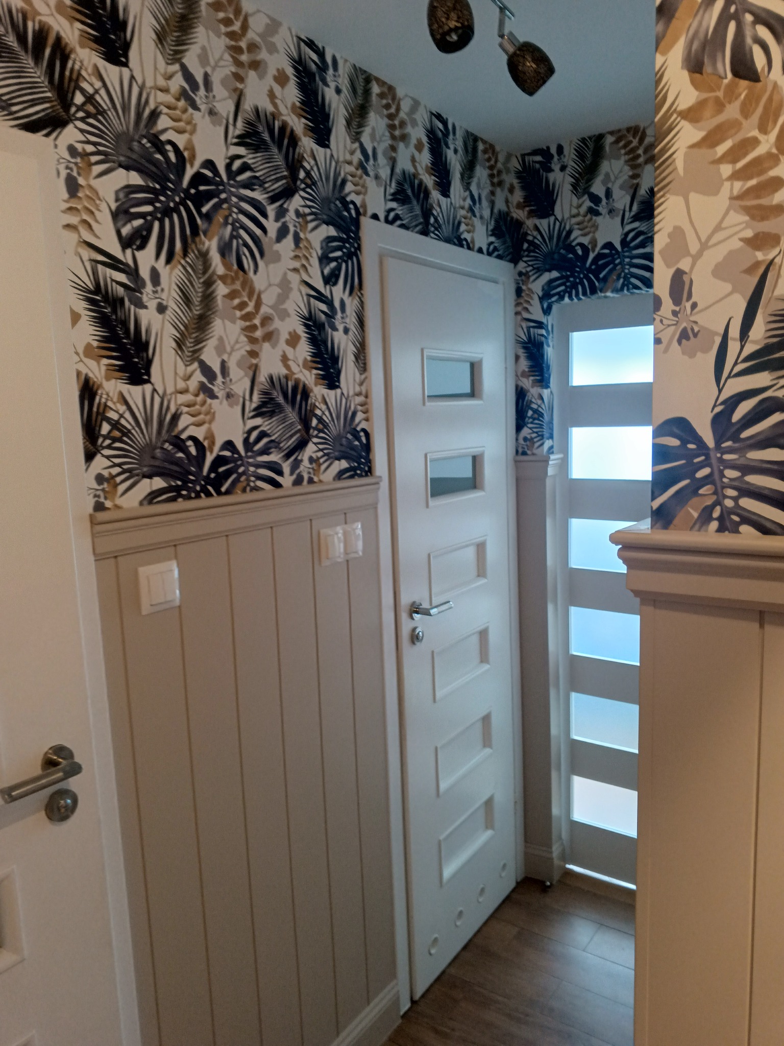 Wnętrze korytarza po remoncie: białe drzwi z przeszkleniami, lamperia w kolorze beżowym i tapeta w motywy roślinne w odcieniach brązu i granatu. Dwa reflektory na suficie.