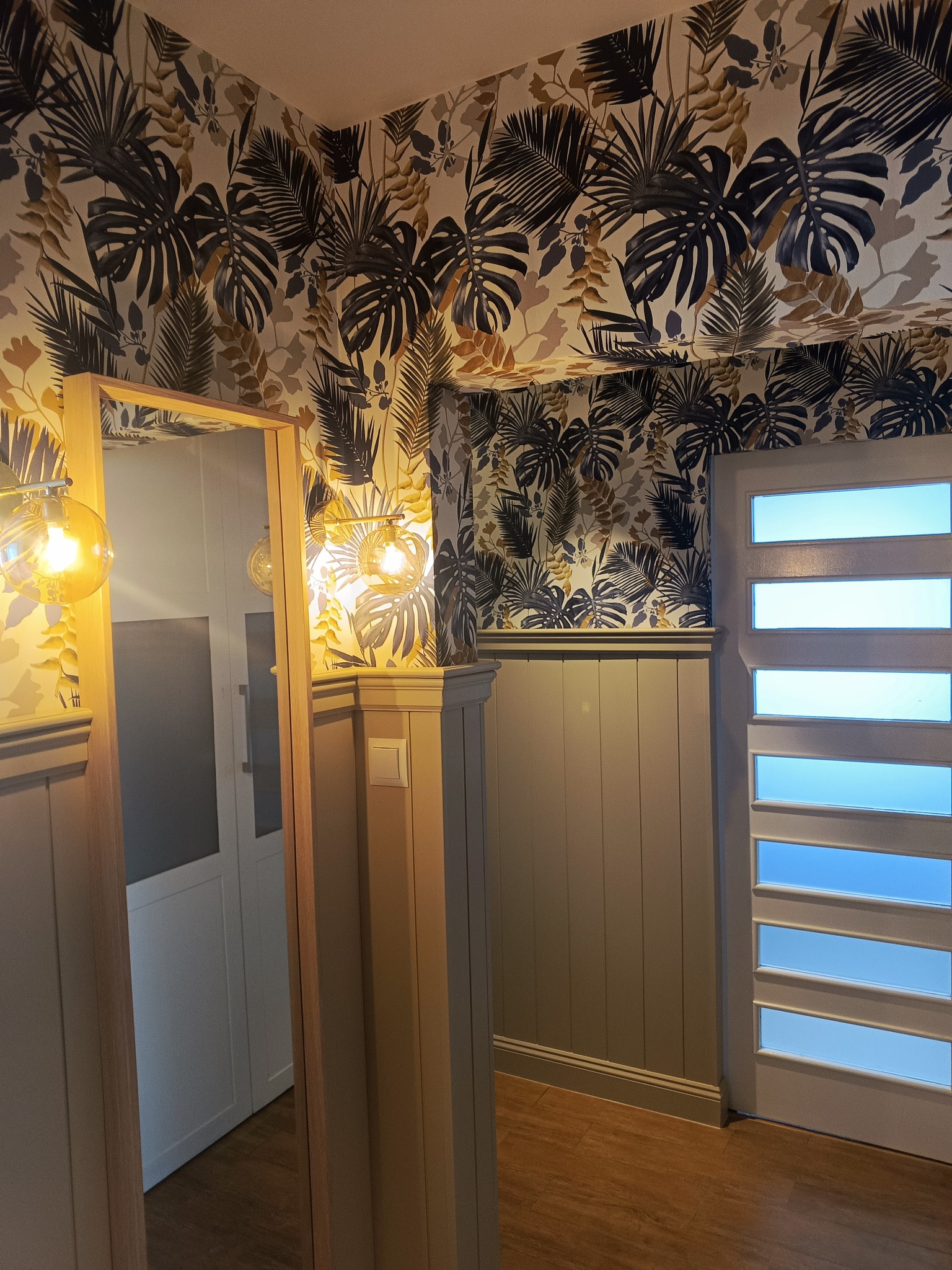 Wykończone wnętrze przedpokoju z tapetą w liście, drewnianą boazerią i lustrem z oświetleniem. Widoczne drzwi z szybami i podłoga z paneli.