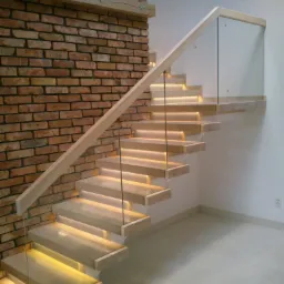 Nowoczesne schody z drewnianymi stopniami podświetlonymi od spodu, szklanymi balustradami i drewnianą poręczą, przylegające do ściany z czerwonej cegły.