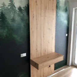 Montaż drewnianej konstrukcji z blatem i szufladą na tle ściany z fototapetą leśną, widoczne przewody elektryczne i listwy przypodłogowe w trakcie montażu.