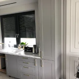 Szara zabudowa kuchenna z frezowanymi frontami, złotymi uchwytami i białym blatem, uzupełniona o akcesoria kuchenne i rośliny doniczkowe na tle okna z roletami.