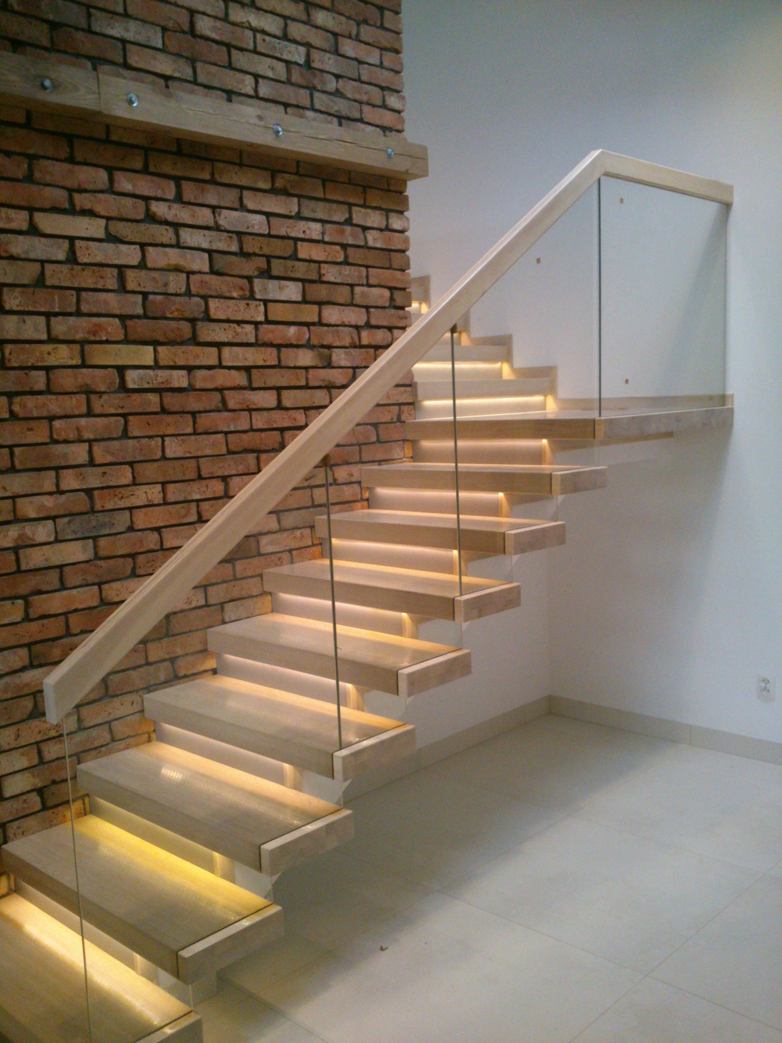 Nowoczesne schody z drewnianymi stopniami podświetlonymi od spodu, szklanymi balustradami i drewnianą poręczą, przylegające do ściany z czerwonej cegły.