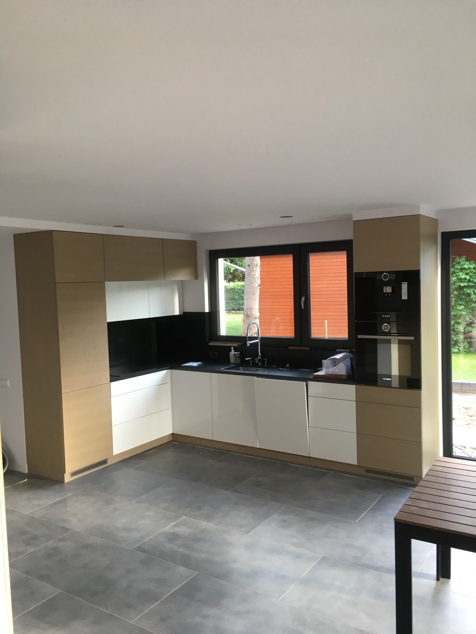 Nowoczesna kuchnia z białymi i drewnopodobnymi frontami, czarnym blatem i zabudowanymi urządzeniami AGD, widok na okno z pomarańczowymi żaluzjami i fragment stołu.