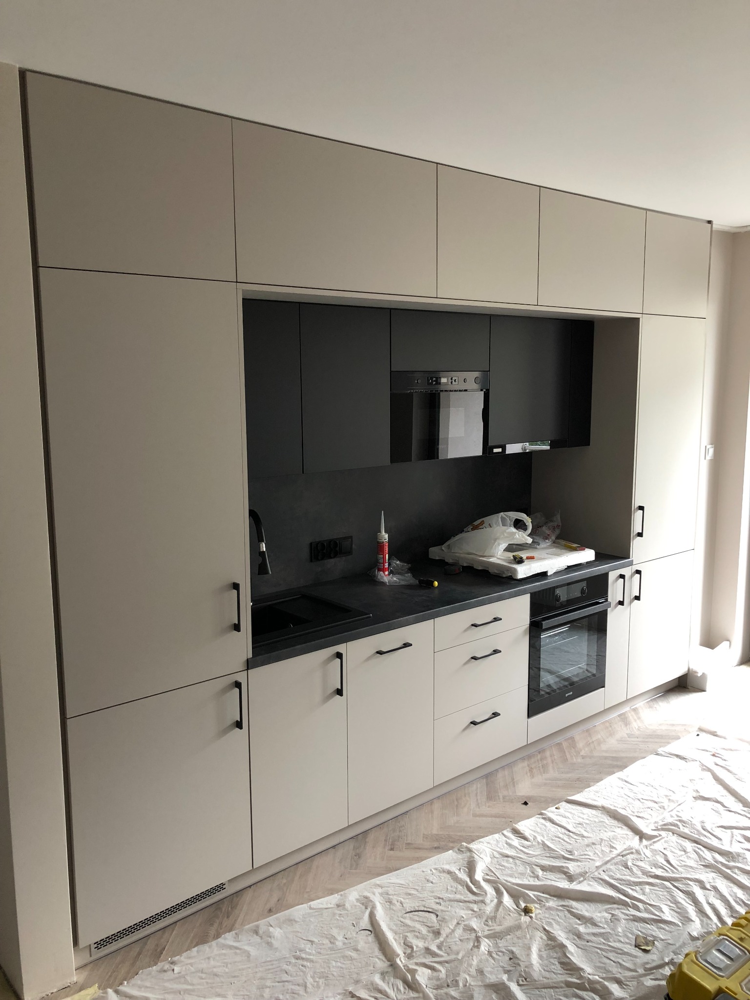 Jasnoszara kuchnia w trakcie montażu, z czarnymi akcentami i czarnym blatem, zabezpieczona folią ochronną na podłodze, widoczny silikon i narzędzia na blacie.