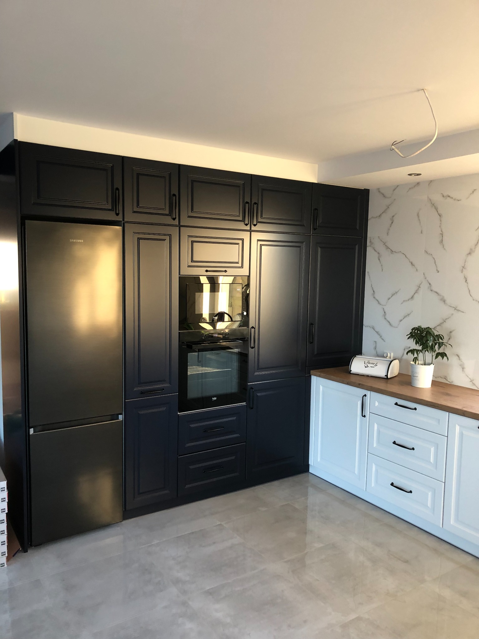 Nowoczesna kuchnia z zabudową w kolorze granatowym, połączona z białymi szafkami z drewnianym blatem. Widoczny sprzęt AGD w zabudowie, w tym lodówka Samsung. Na blacie stoi biały chlebak i doniczka...