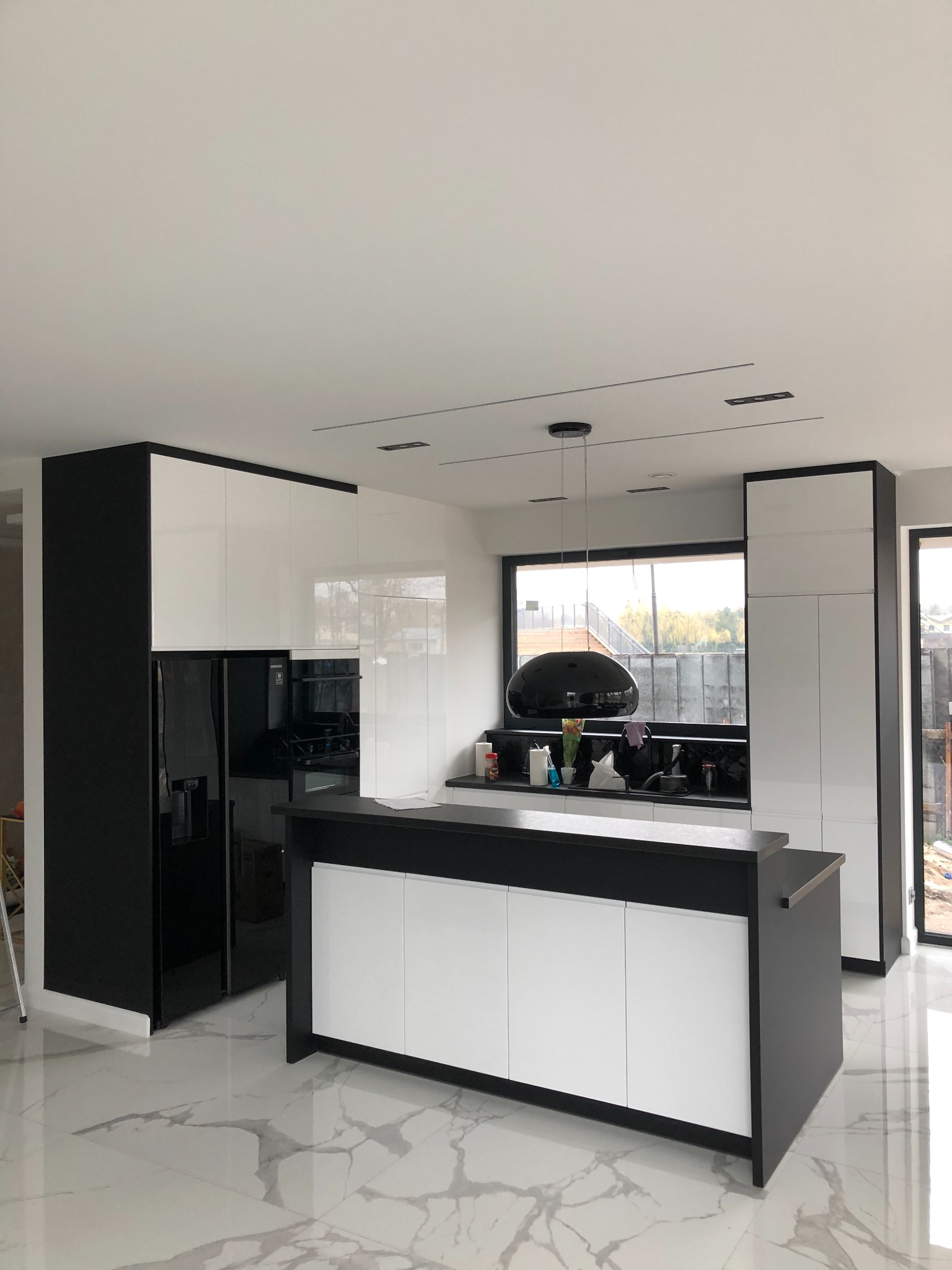 Nowoczesna, minimalistyczna kuchnia z wyspą, czarno-białe szafki z połyskiem, marmurowa podłoga, czarna lampa wisząca, widok na zewnątrz przez okno.