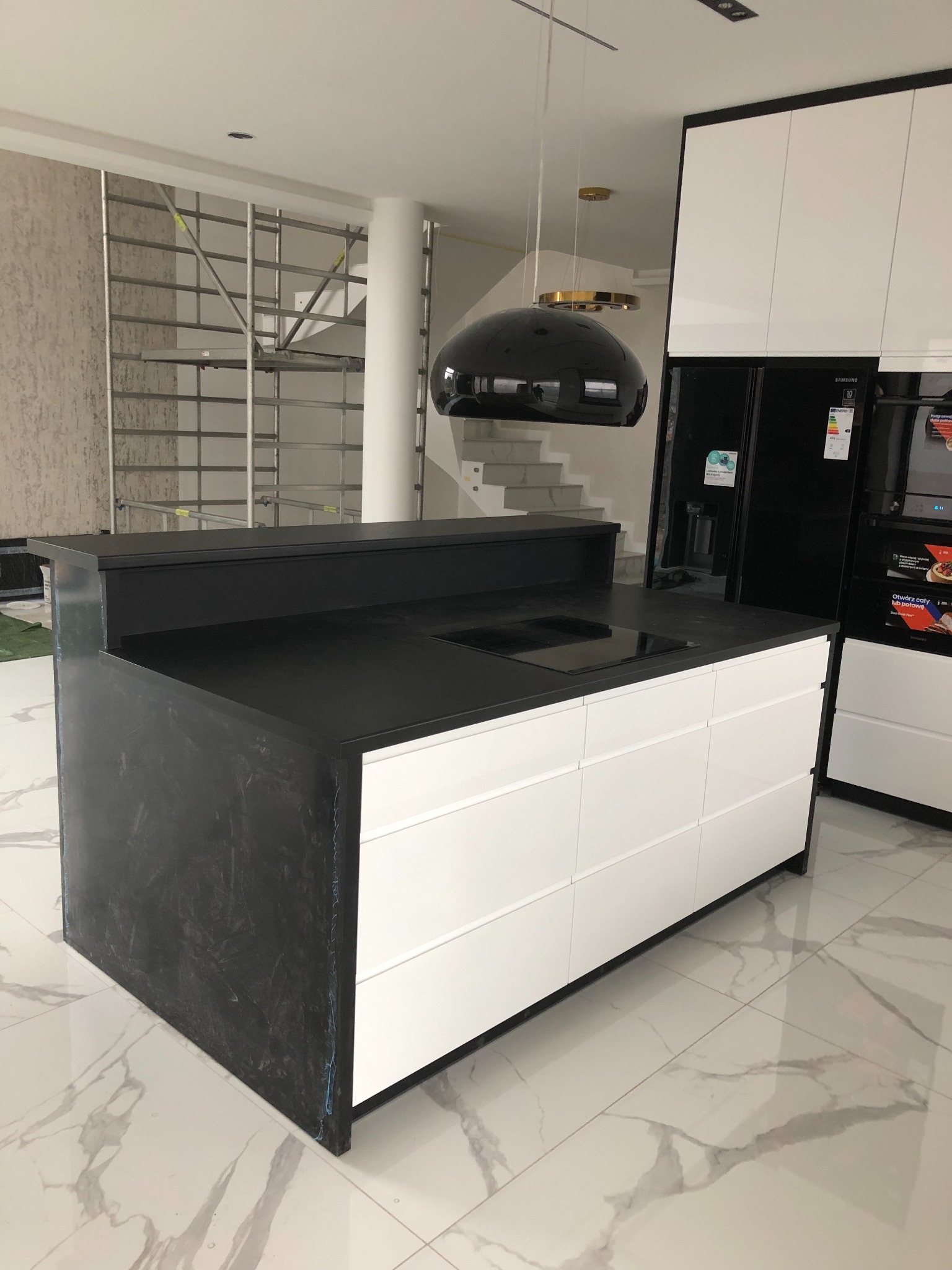 Wyspa kuchenna z czarnym blatem i białymi frontami szuflad, widoczna płyta indukcyjna, w tle rusztowanie i schody, podwieszana czarna lampa.