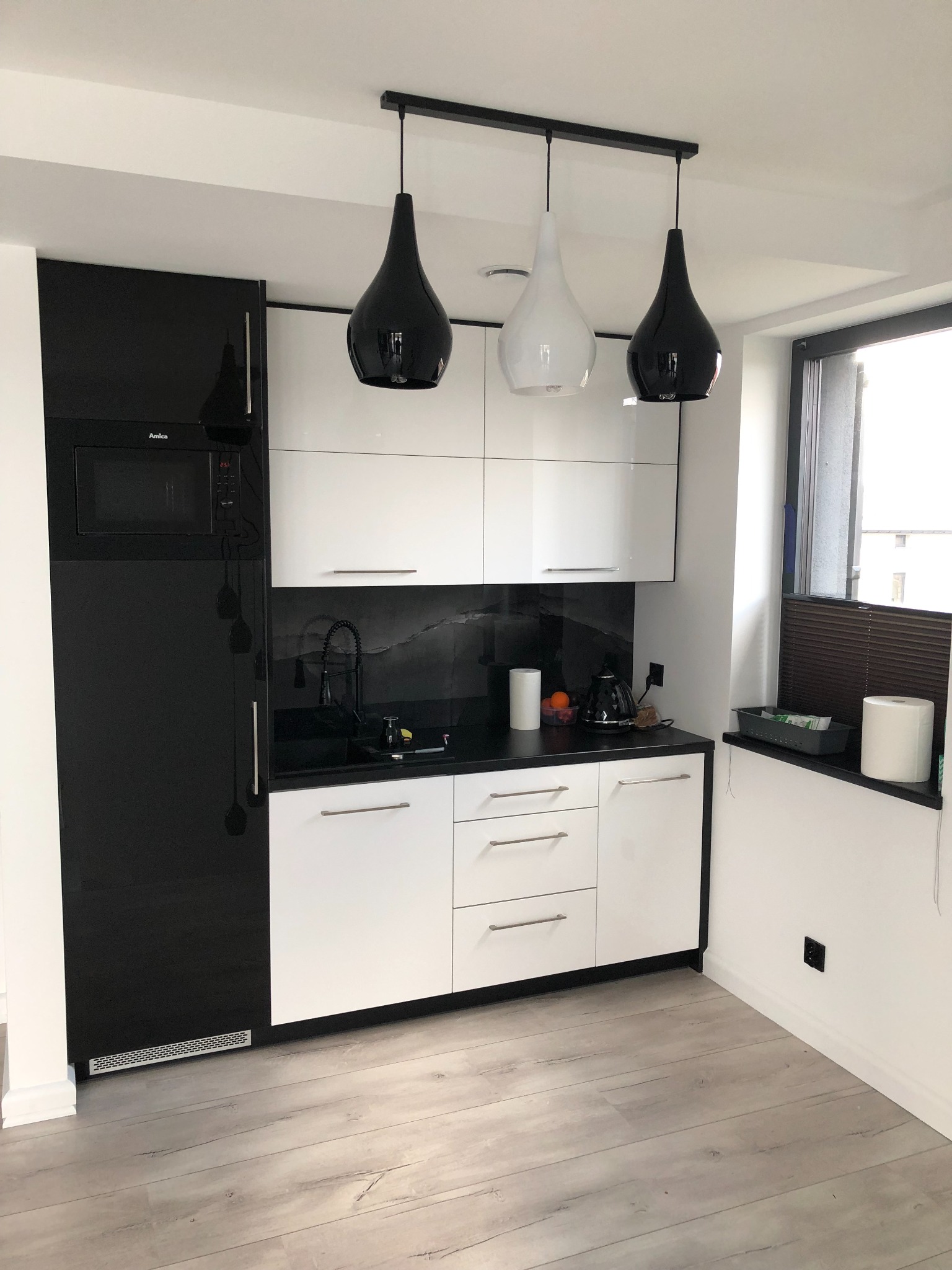 Nowoczesna kuchnia z czarno-białymi szafkami, czarnym blatem, trzema wiszącymi lampami w kształcie łezki (dwie czarne, jedna biała) i mikrofala Amica w zabudowie.