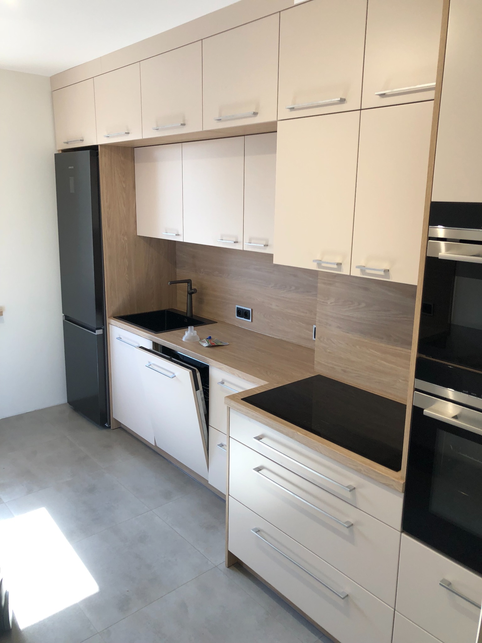 Nowoczesna kuchnia z jasnymi frontami szafek, czarnym zlewem i płytą indukcyjną, oraz zabudowaną lodówką, z otwartą zmywarką i drobnymi przedmiotami na blacie.