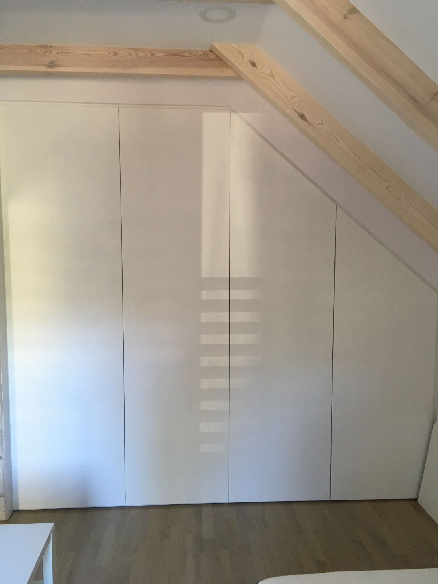 Biała zabudowa wnękowa pod skosem na poddaszu, widoczne drewniane belki stropowe i parkiet na podłodze.