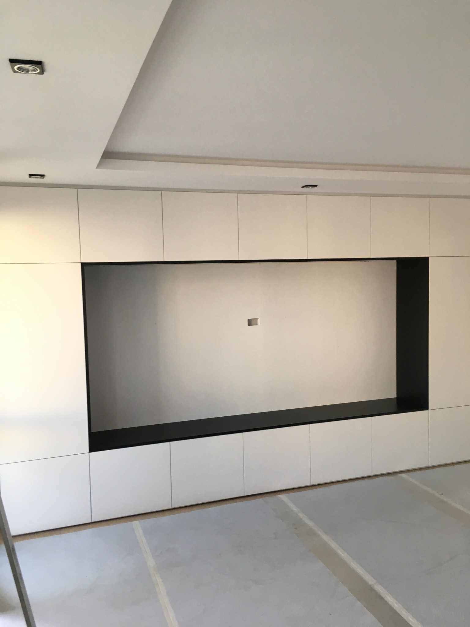 Biała zabudowa meblowa z czarną wnęką na ścianie, widoczne oświetlenie sufitowe i fragment podłogi w trakcie prac wykończeniowych.