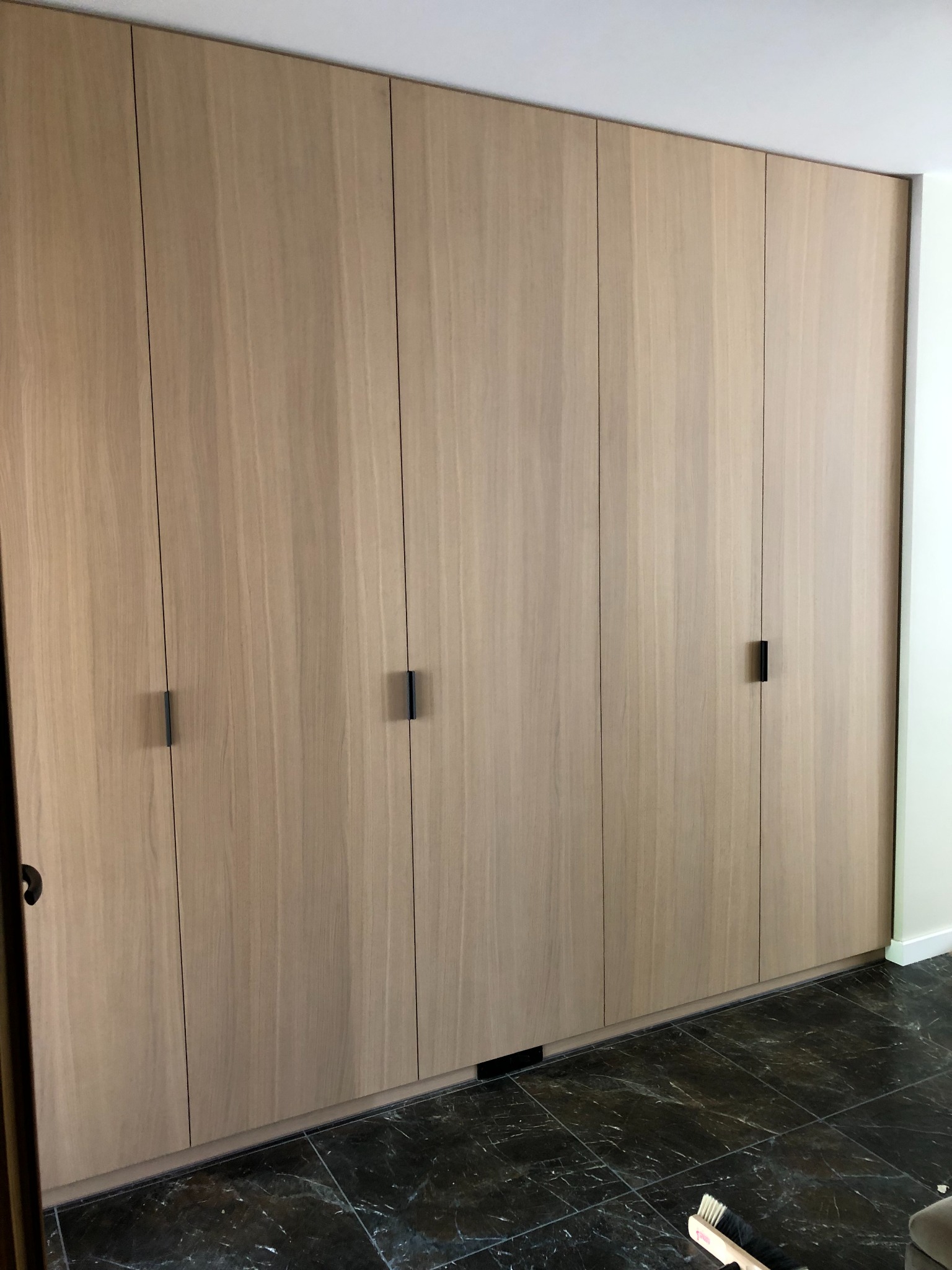 Szafa wnękowa z jasnego drewna z minimalistycznymi, czarnymi uchwytami, dopasowana do sufitu, na podłodze z ciemnego marmuru, widoczny fragment szczotki do zamiatania.