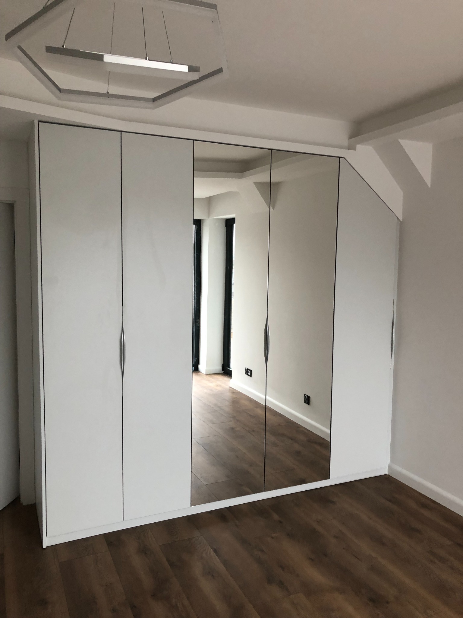 Biała szafa wnękowa z lustrzanymi drzwiami, dopasowana do skosu sufitu, na podłodze panele w odcieniach brązu, widoczne gniazdka elektryczne.