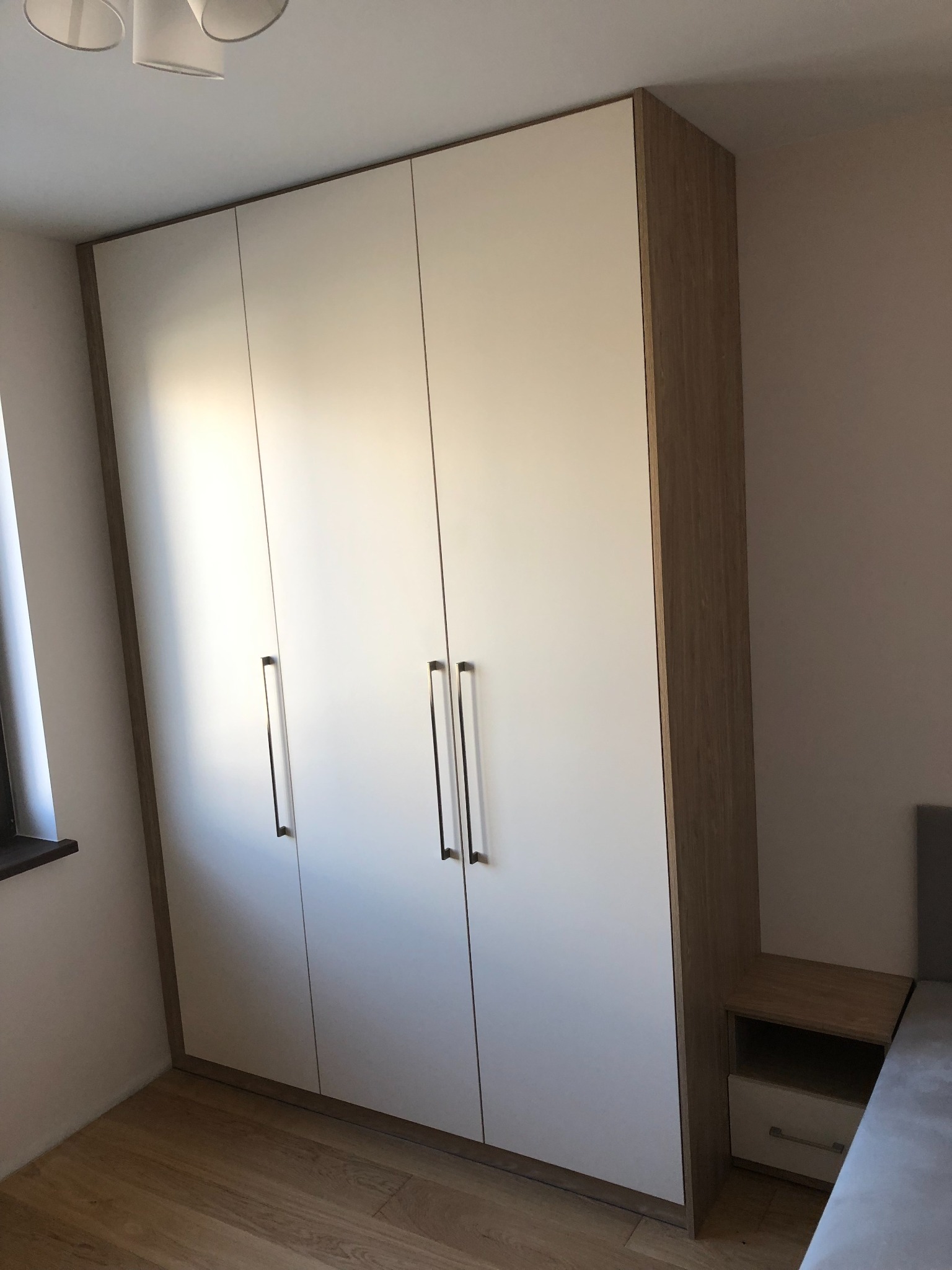 Trzydrzwiowa szafa z białymi frontami i obramowaniem w kolorze jasnego drewna, dopasowana do szafki nocnej obok łóżka.