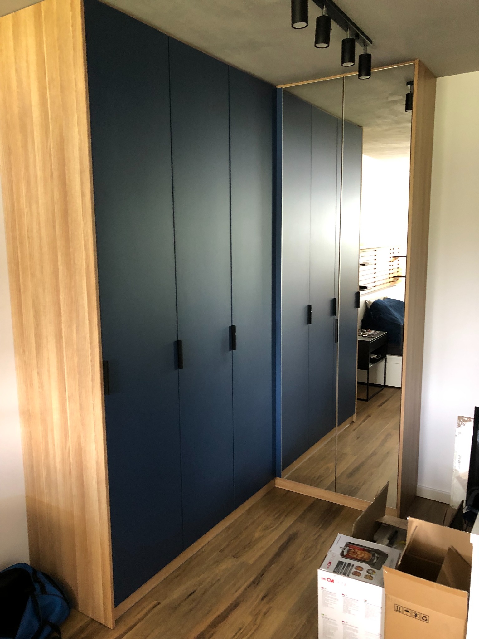 Nowoczesna szafa wnękowa z ciemnoniebieskimi frontami i drewnianymi bokami, z lustrzanymi drzwiami po prawej stronie, odbijającymi wnętrze pokoju.
