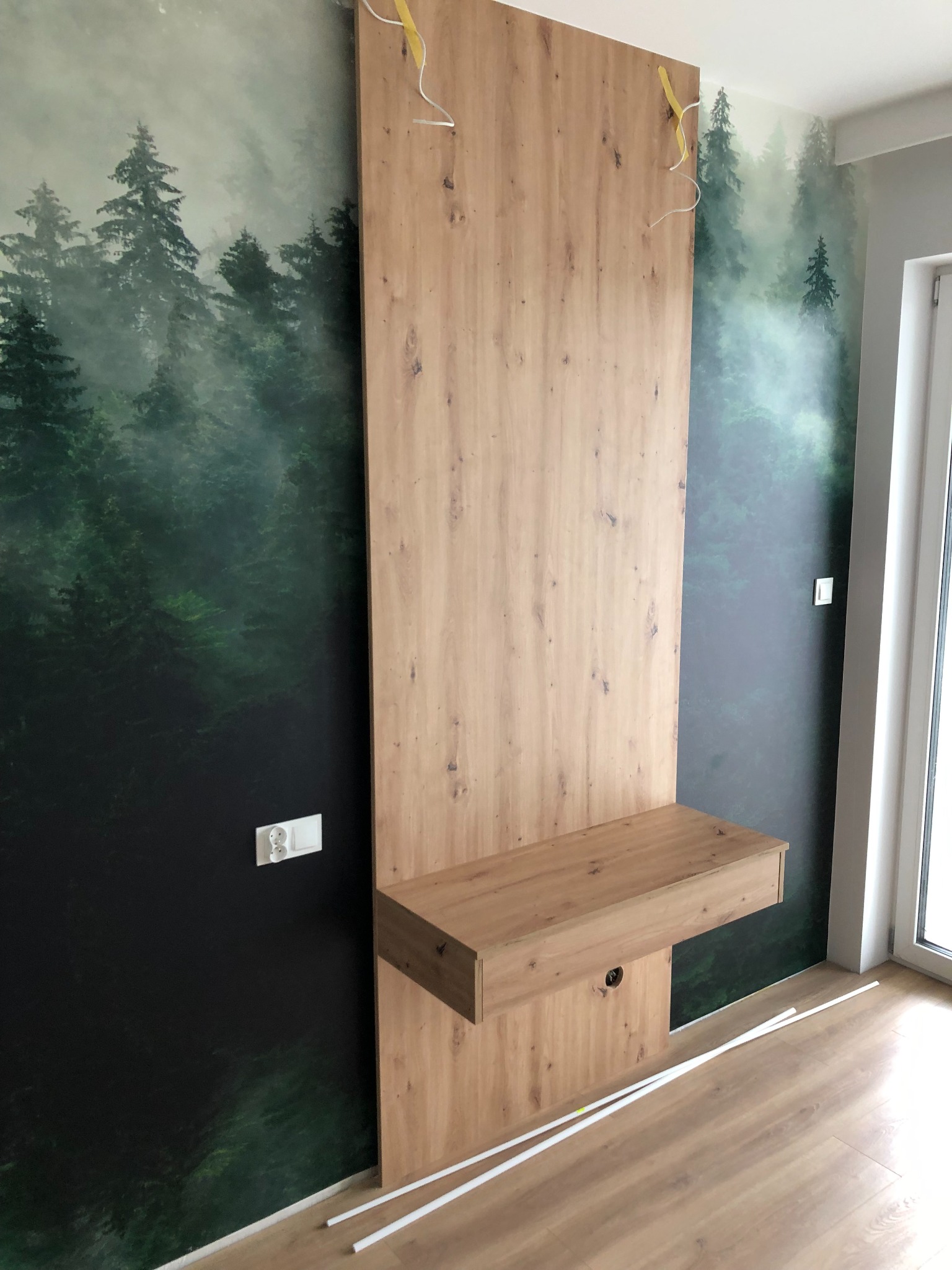 Montaż drewnianej konstrukcji z blatem i szufladą na tle ściany z fototapetą leśną, widoczne przewody elektryczne i listwy przypodłogowe w trakcie montażu.