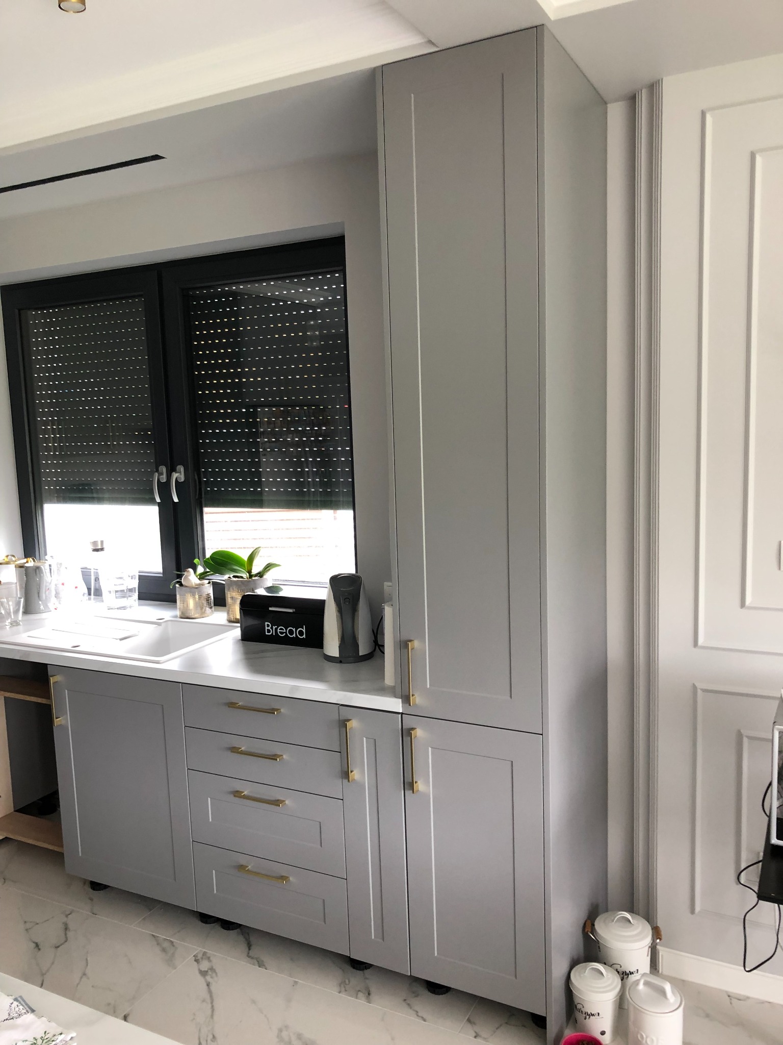 Szara zabudowa kuchenna z frezowanymi frontami, złotymi uchwytami i białym blatem, uzupełniona o akcesoria kuchenne i rośliny doniczkowe na tle okna z roletami.