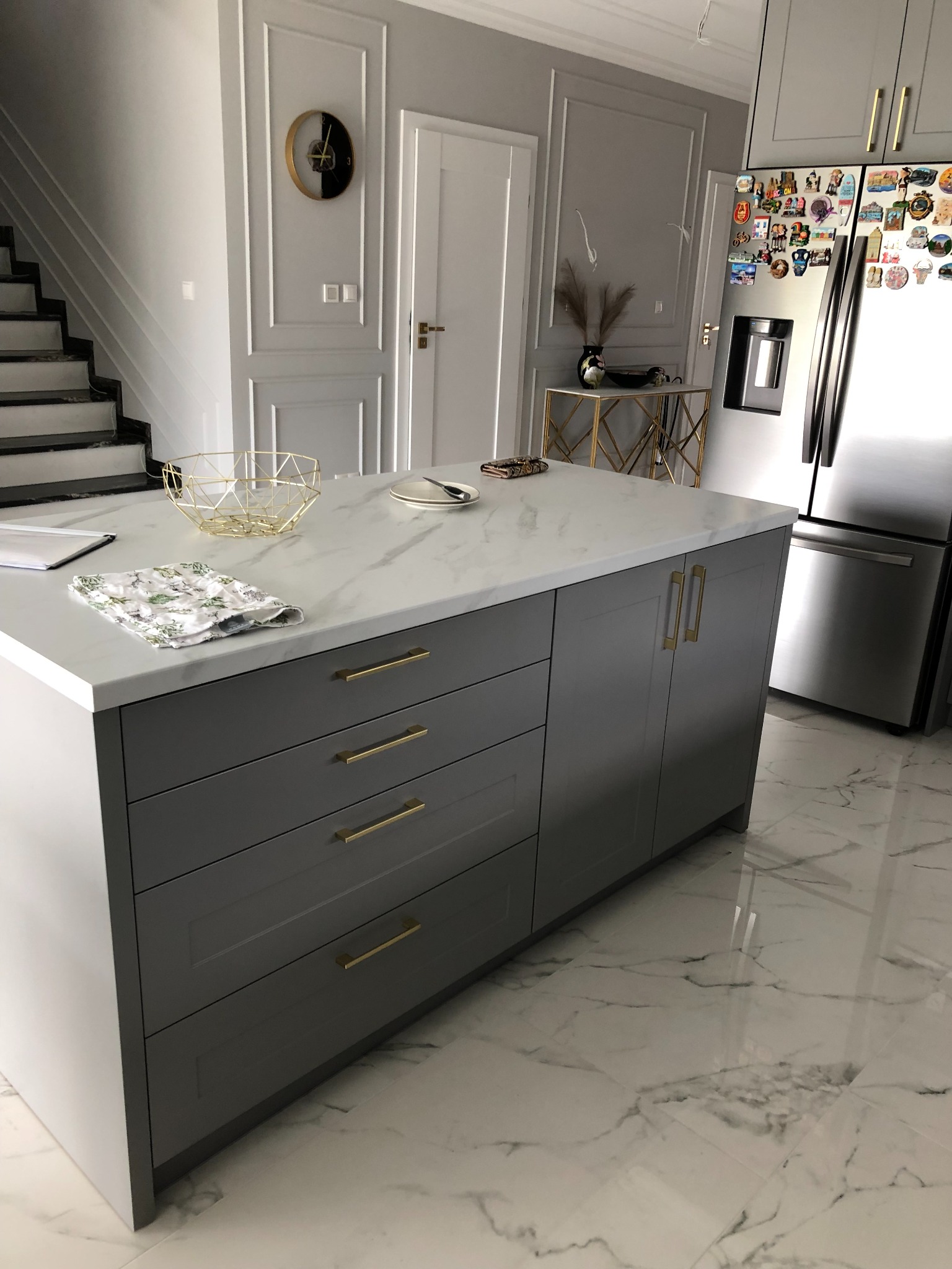 Szara wyspa kuchenna z marmurowym blatem, złotymi uchwytami i elementami dekoracyjnymi, na marmurowej posadzce, w tle schody i lodówka.