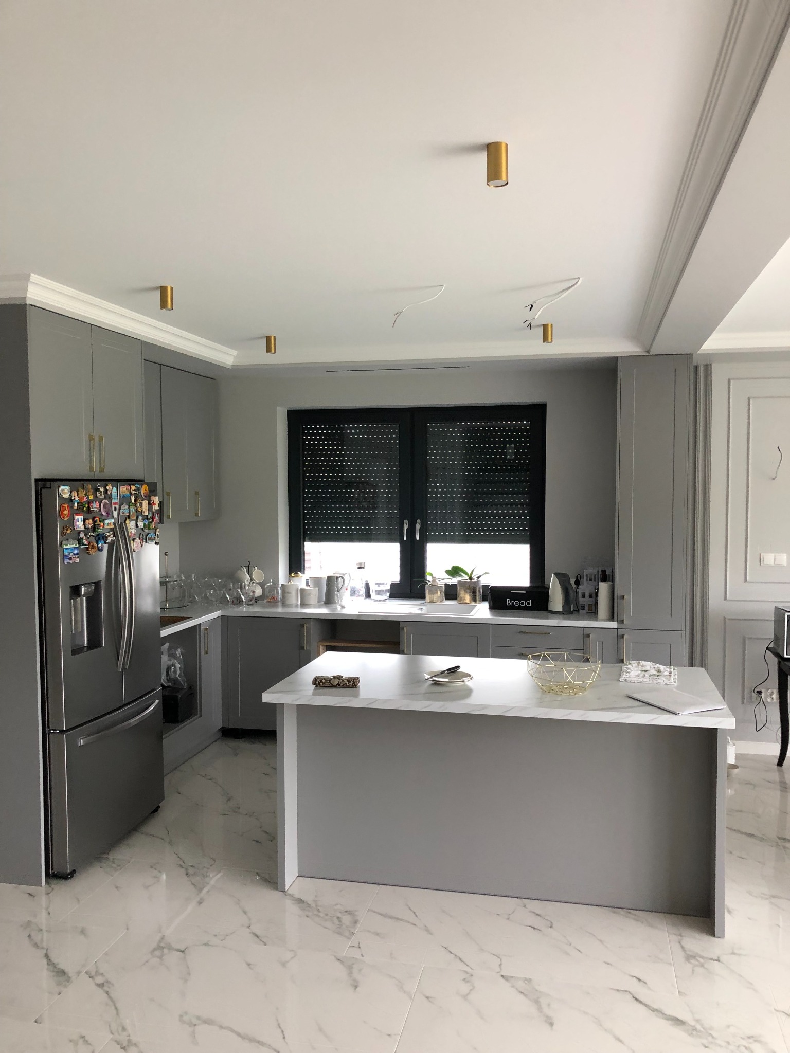 Nowoczesna kuchnia w odcieniach szarości z marmurową podłogą i wyspą kuchenną, złote akcenty w oświetleniu i uchwytach, lodówka obklejona magnesami, okno z roletami.