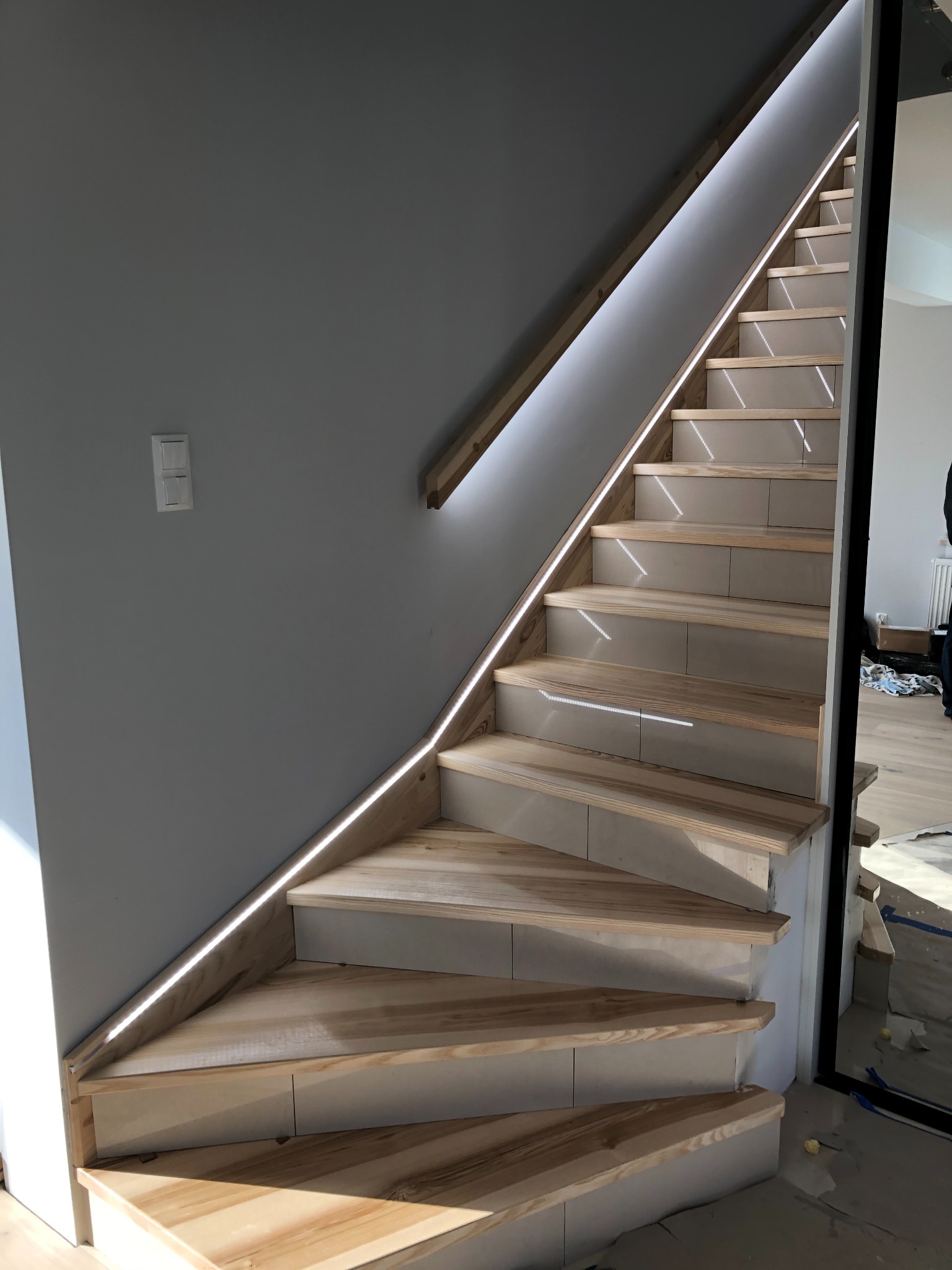 Nowoczesne schody drewniane z podświetleniem LED wzdłuż ściany, widok z dołu, jasne drewno, szara ściana, minimalistyczny design.