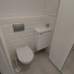 Zdrojewsky - Nowoczesna, całkowicie biała łazienka z podwieszaną toaletą, umywalką z szafką i lustrem w zabudowie. Ściany wyłożone białymi, prostokątnymi płytkami imitującymi cegłę, szara podłoga z płytek.