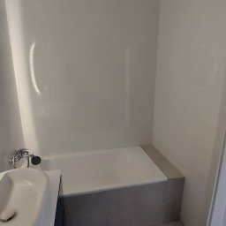 Zdrojewsky - Nowoczesna łazienka z wanną obudowaną szarą płytką, białą armaturą i panelami ściennymi.