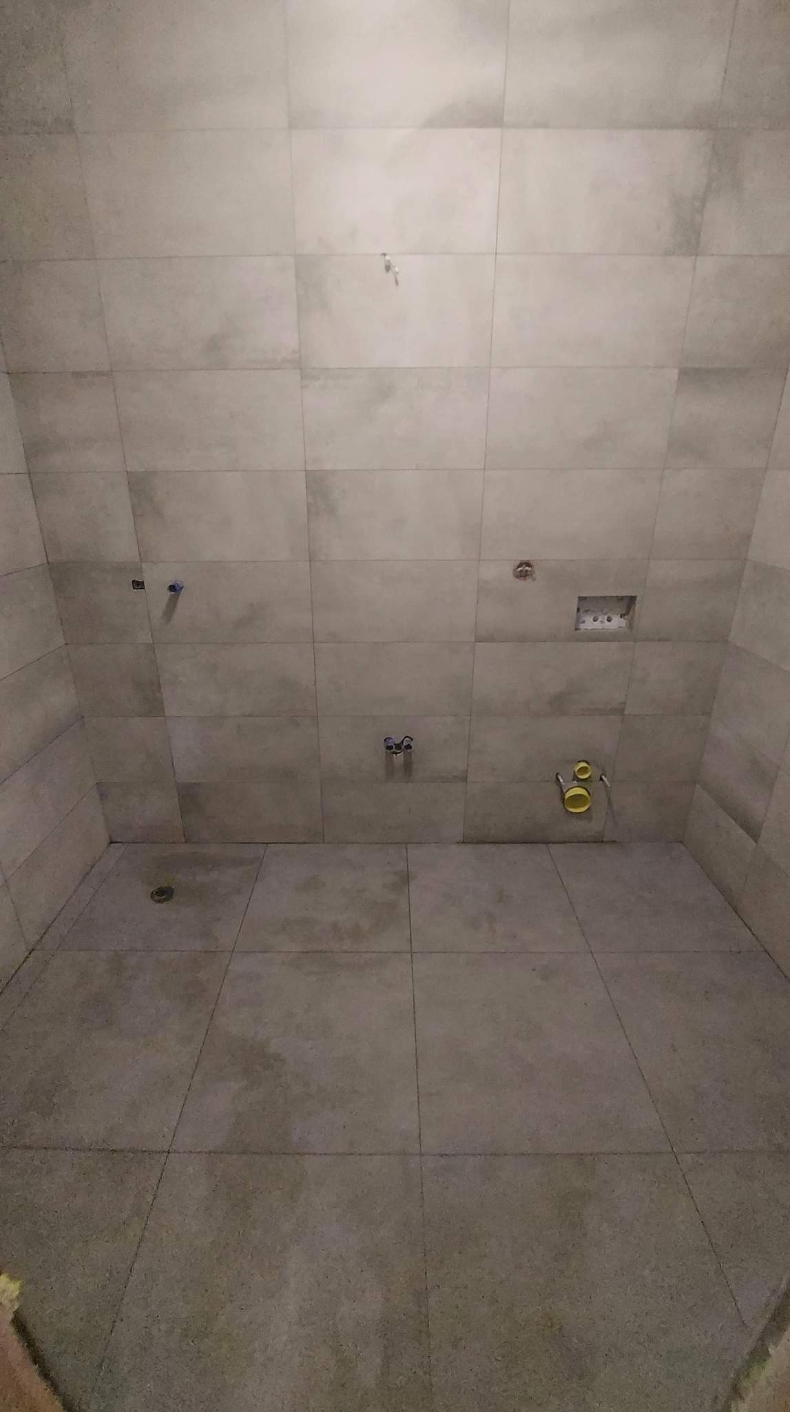 Wnętrze łazienki w trakcie remontu, wyłożone płytkami w odcieniach szarości, z widocznymi instalacjami hydraulicznymi przed montażem armatury.