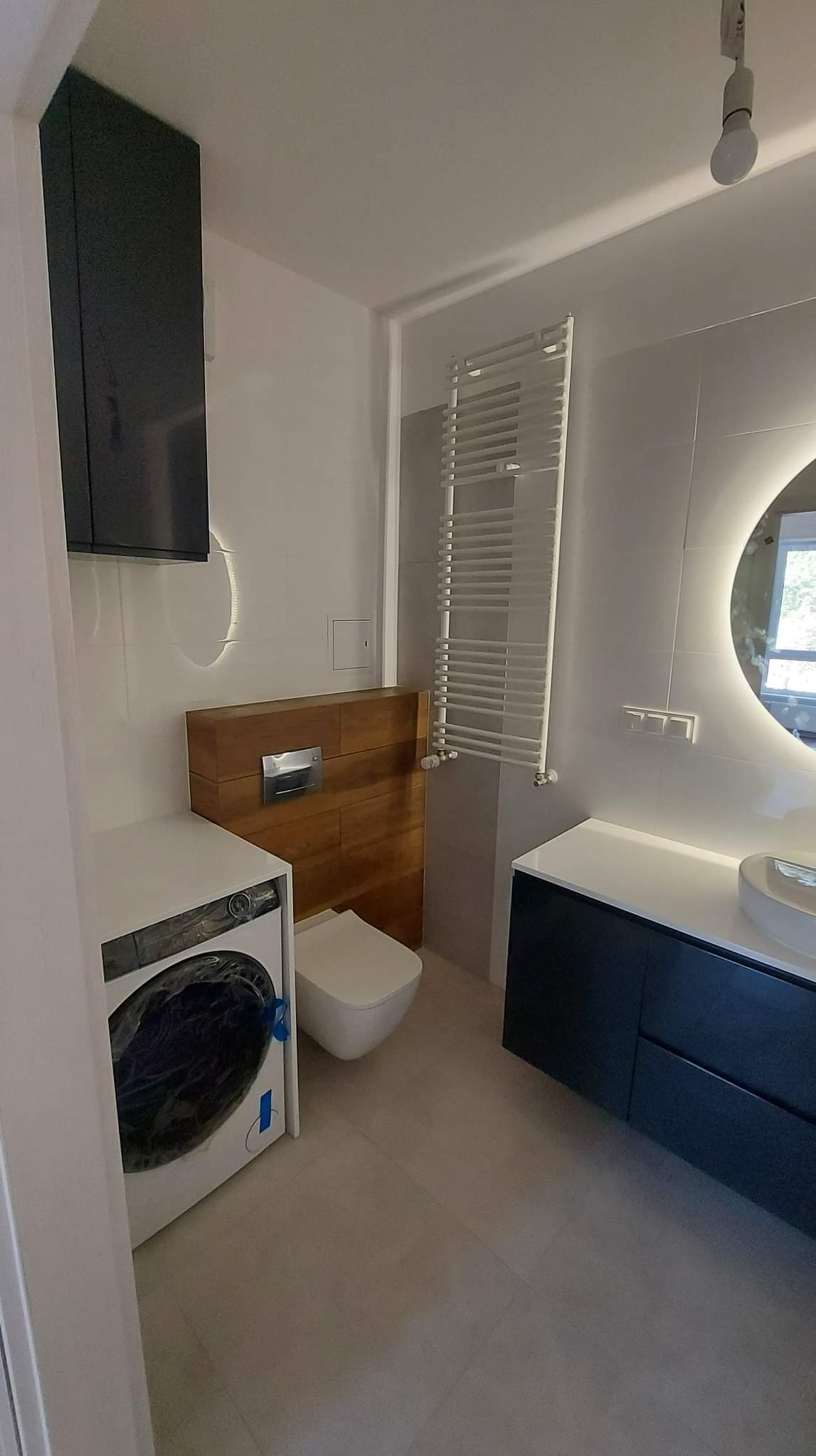 Nowoczesna łazienka z pralką, podwieszaną toaletą z drewnianą obudową, grzejnikiem drabinkowym i okrągłym lustrem z podświetleniem LED.