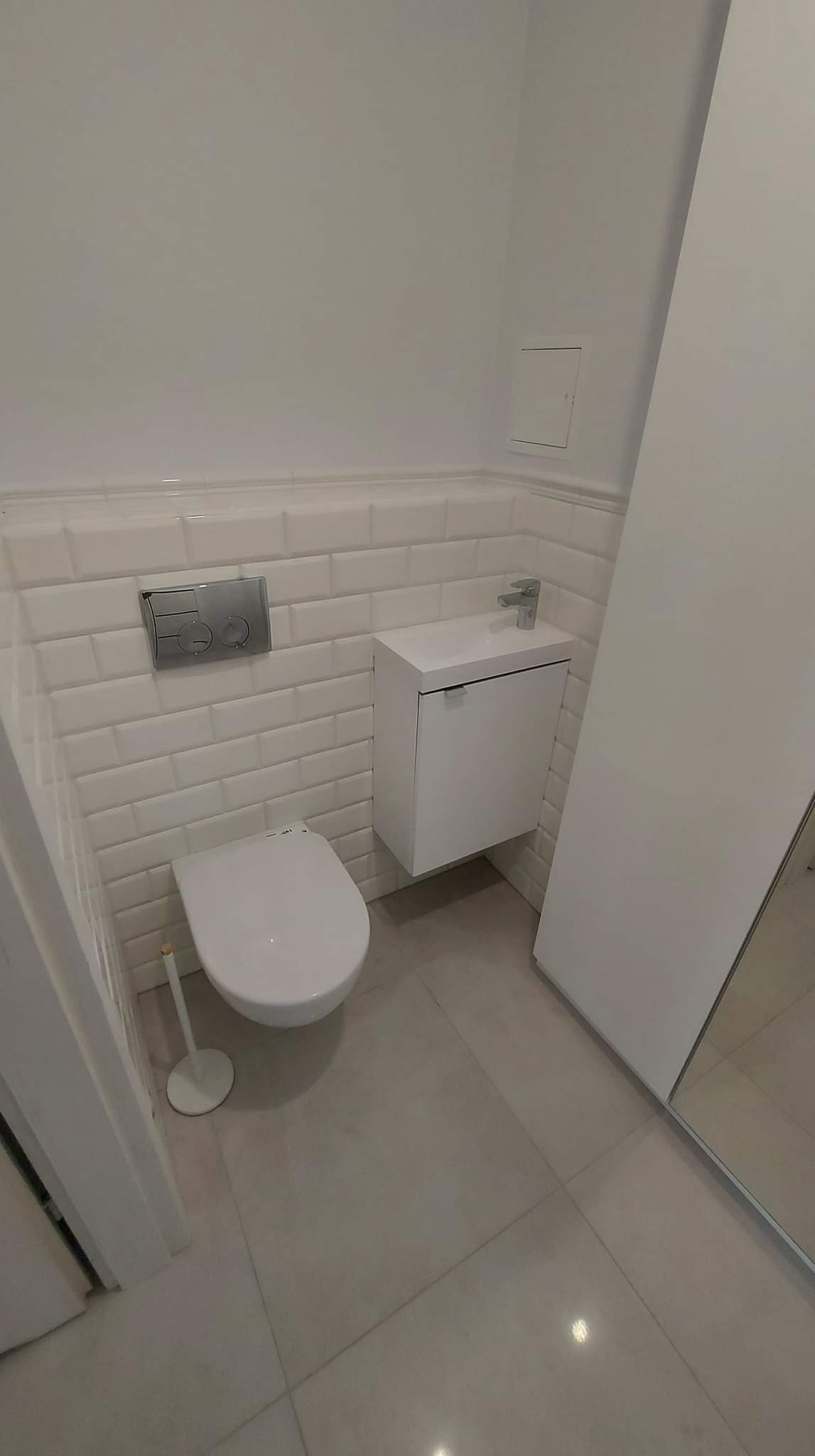 Nowoczesna, całkowicie biała łazienka z podwieszaną toaletą, umywalką z szafką i lustrem w zabudowie. Ściany wyłożone białymi, prostokątnymi płytkami imitującymi cegłę, szara podłoga z płytek.