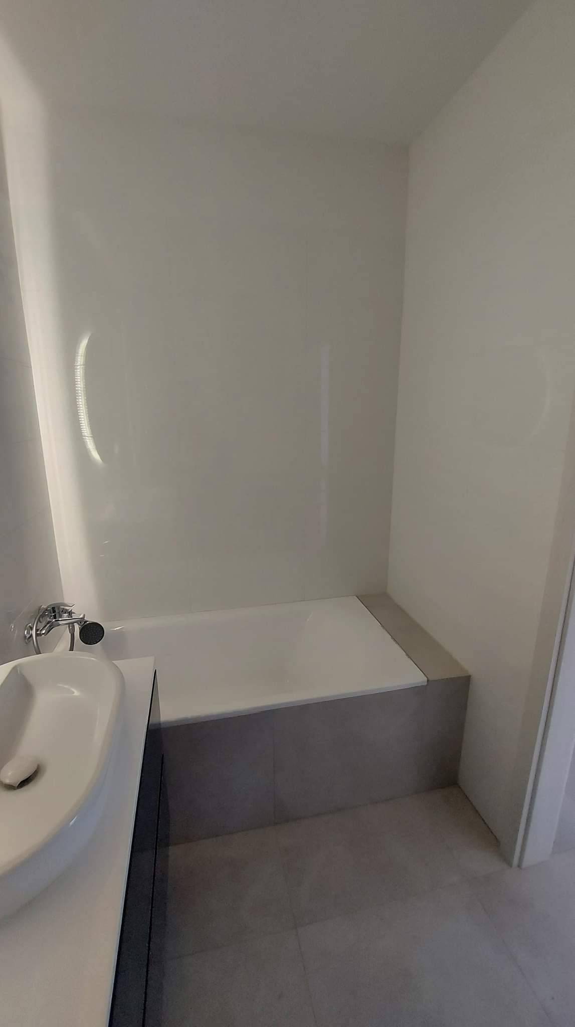 Nowoczesna łazienka z wanną obudowaną szarą płytką, białą armaturą i panelami ściennymi.