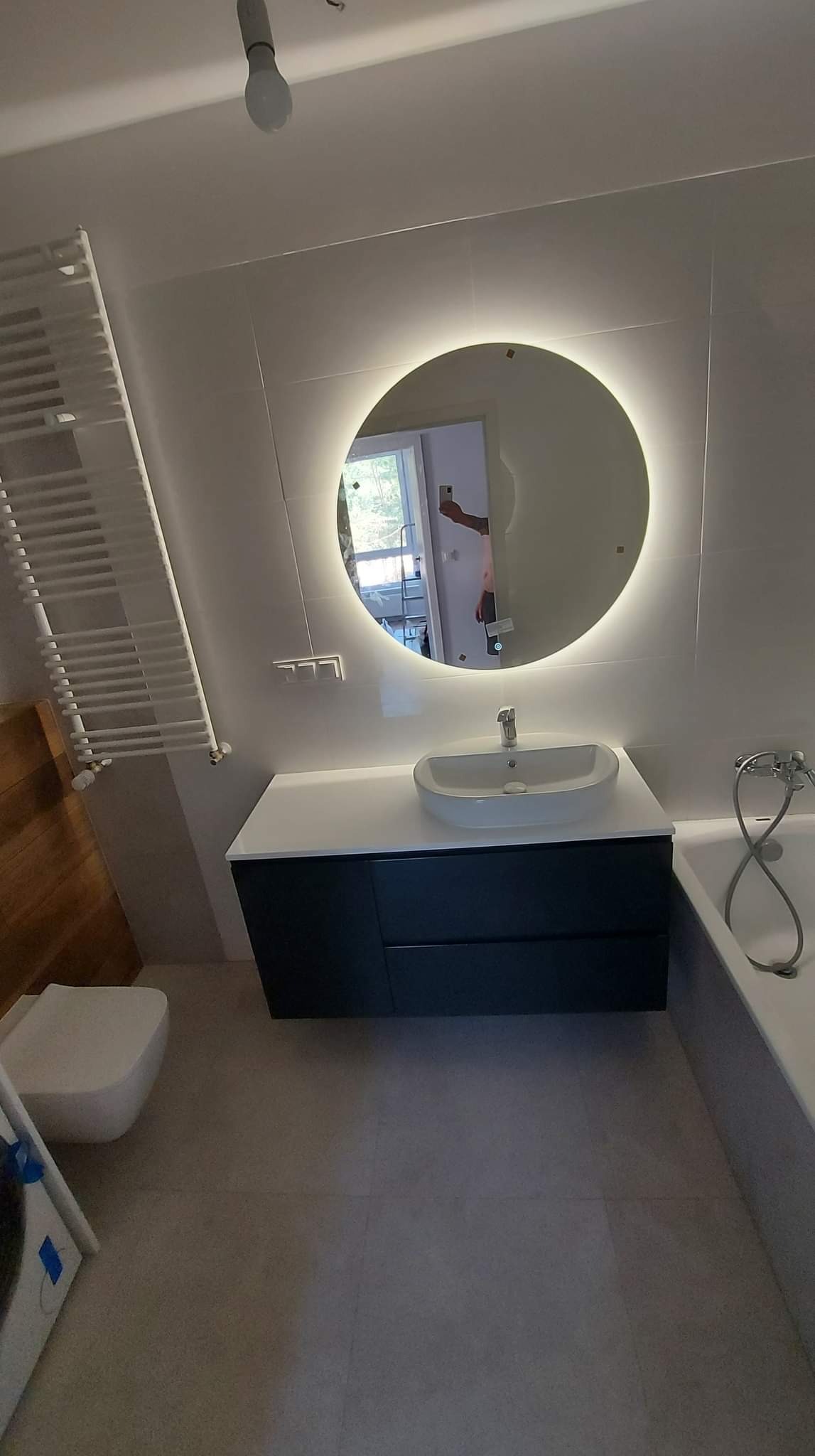 Nowoczesna łazienka z okrągłym lustrem podświetlanym LED, umywalką nablatową na szafce z ciemnymi frontami oraz grzejnikiem drabinkowym.