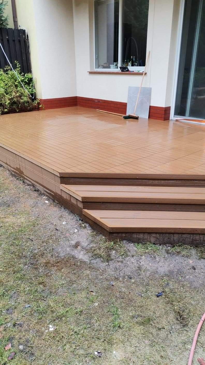 Nowy taras z kompozytu drewnopodobnego w kolorze brązowym, z trzema schodkami, przylegający do budynku z oknem i ceglaną podmurówką, widok z poziomu trawnika.