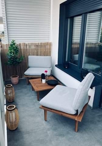 Przytulny balkon z szarymi płytkami podłogowymi, drewnianymi meblami ogrodowymi z jasnymi poduszkami, bambusową osłoną i roletami zewnętrznymi.