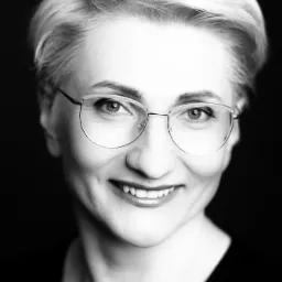 Portret uśmiechniętej kobiety w okularach z krótkimi blond włosami na czarnym tle.