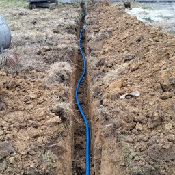USŁUGI INSTALACYJNE PAWLED - Niebieski przewód w wykopie ziemnym, widoczny na tle minikoparki i zabudowań. Instalacja podziemna w trakcie realizacji.