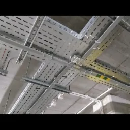 Metalowe korytka kablowe, perforowane, zamocowane pod sufitem. Widoczne białe rury instalacyjne. Szare, betonowe tło sufitu. Kąt ujęcia od dołu.