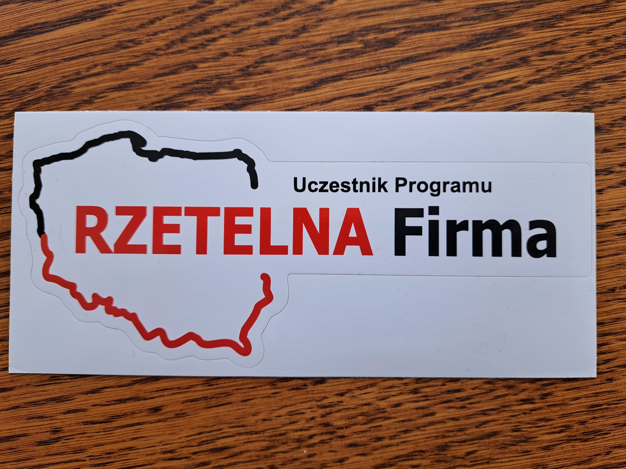 Naklejka 'Uczestnik Programu Rzetelna Firma' z obrysem Polski w kolorach czerni i czerwieni, leżąca na drewnianym tle.