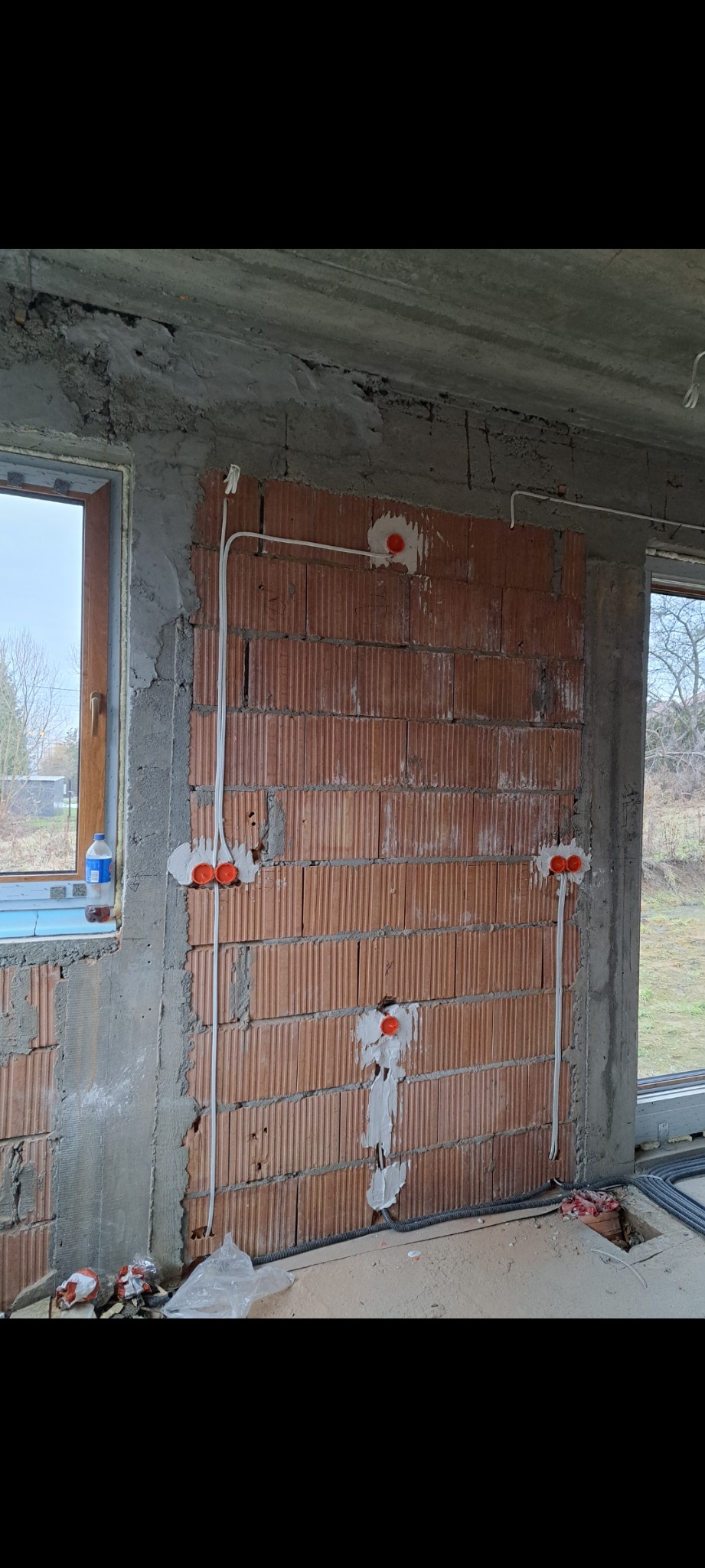 Ceglana ściana w surowym stanie z zamontowanymi puszkami i przewodami elektrycznymi. Widoczne okno i fragment podłogi z przewodami w peszlu.