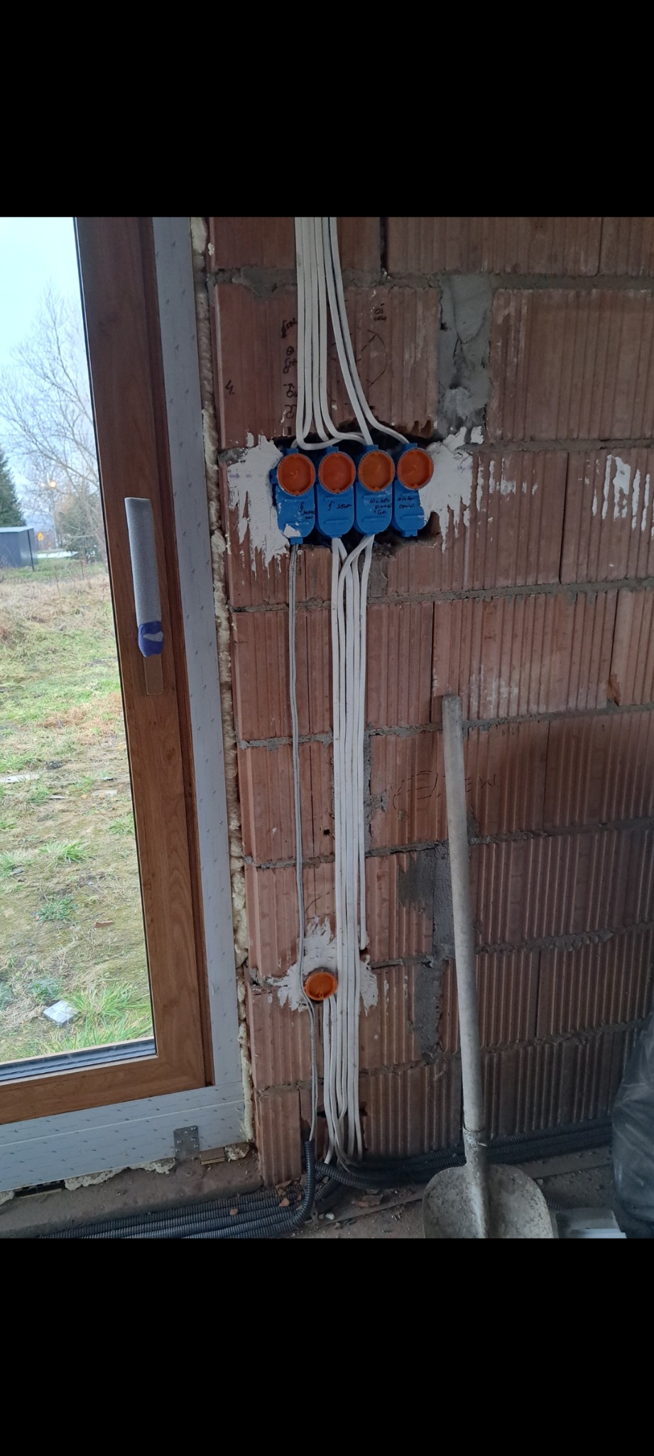 Wewnętrzna instalacja elektryczna w surowym murze, widoczne przewody w peszlach i puszki instalacyjne. Okno z widokiem na zewnątrz. Etap prac przed tynkowaniem.